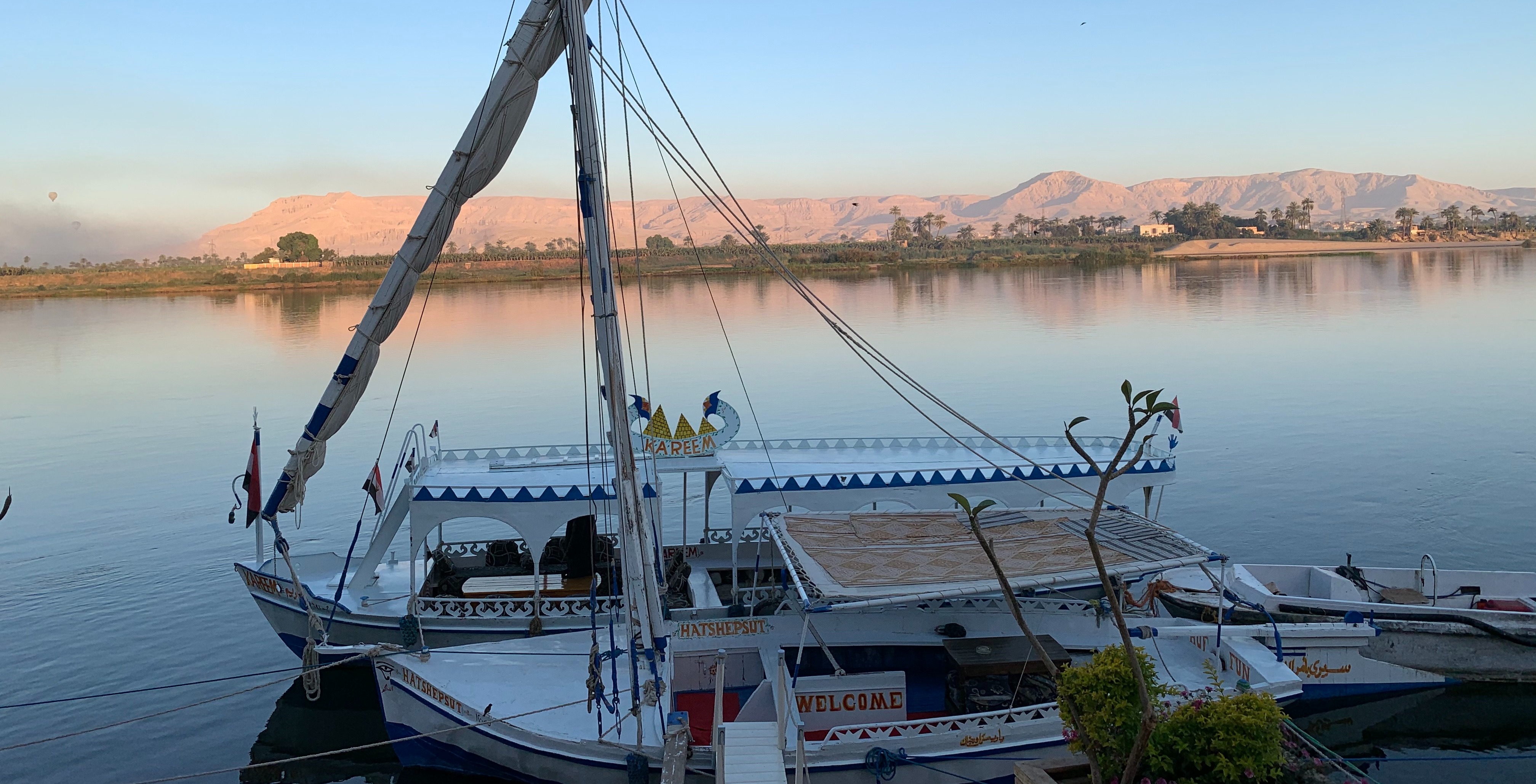 The image features a traditional Egyptian felucca sailing on a tranquil river with mountains in the backdrop during a serene time of day.