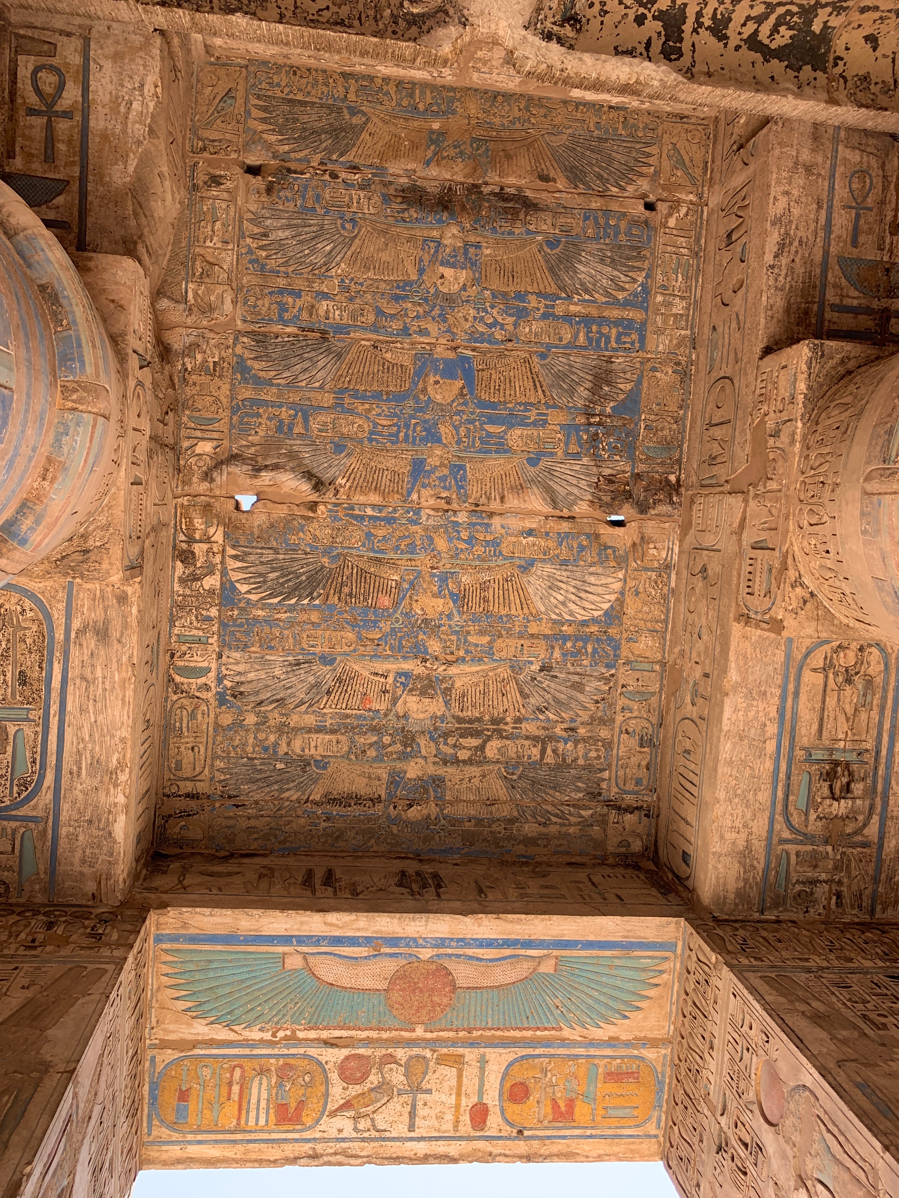 The image depicts the ornate ceiling of an ancient Egyptian structure adorned with hieroglyphics and symbolic art in blue, ochre, and white hues.