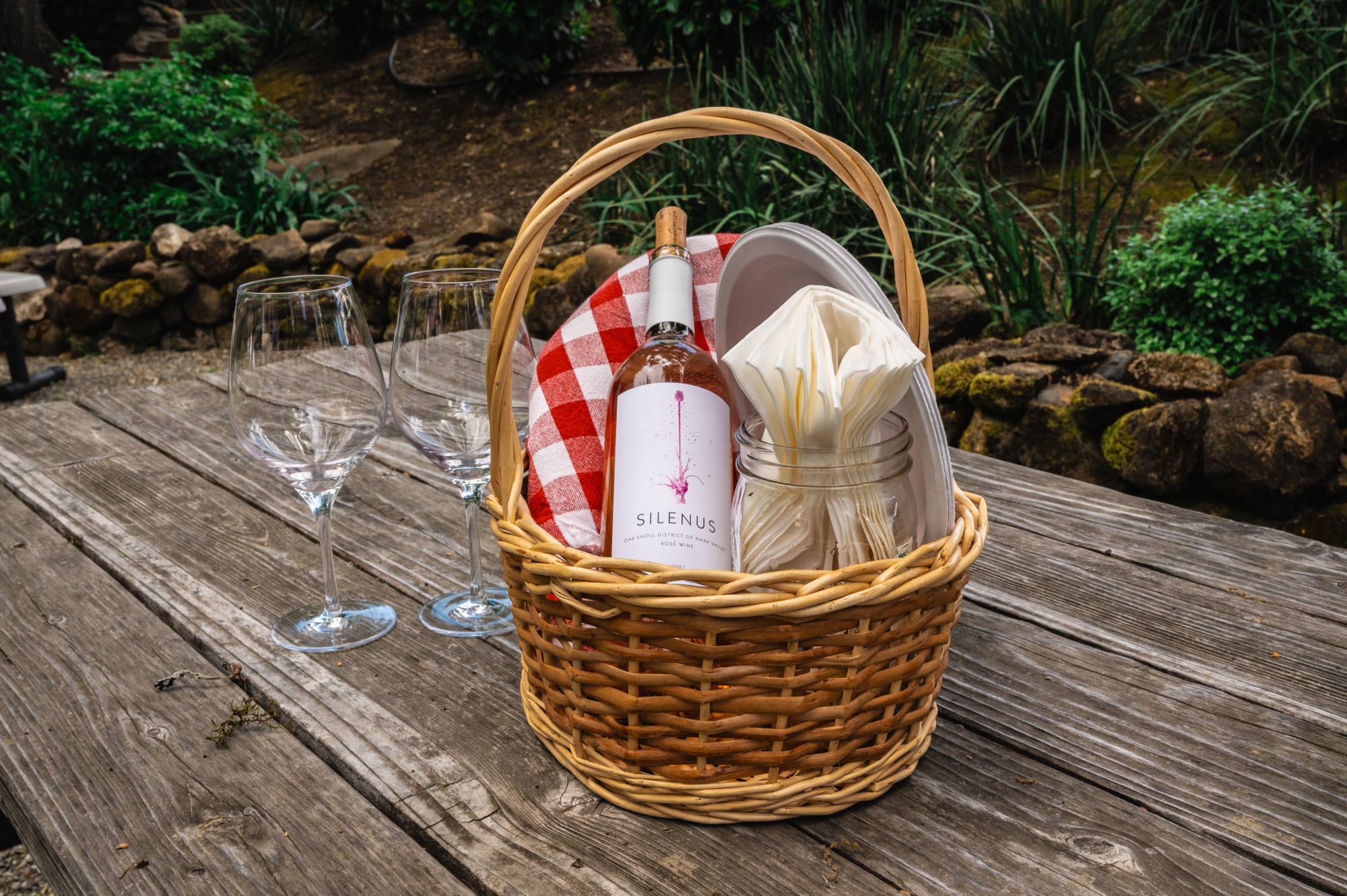 A Silenus wine bottle in a picnic basket next to two glasses on a wooden picnic table.