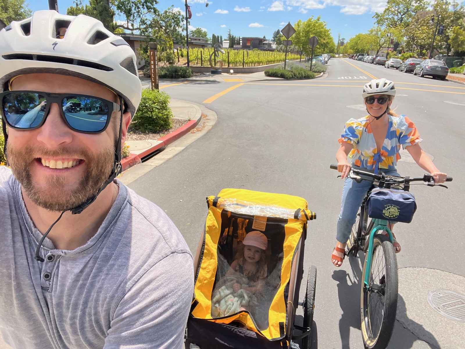 A selfie of Chelsea and her partner biking, with their daughter in tow in a yellow attachment on wheels.