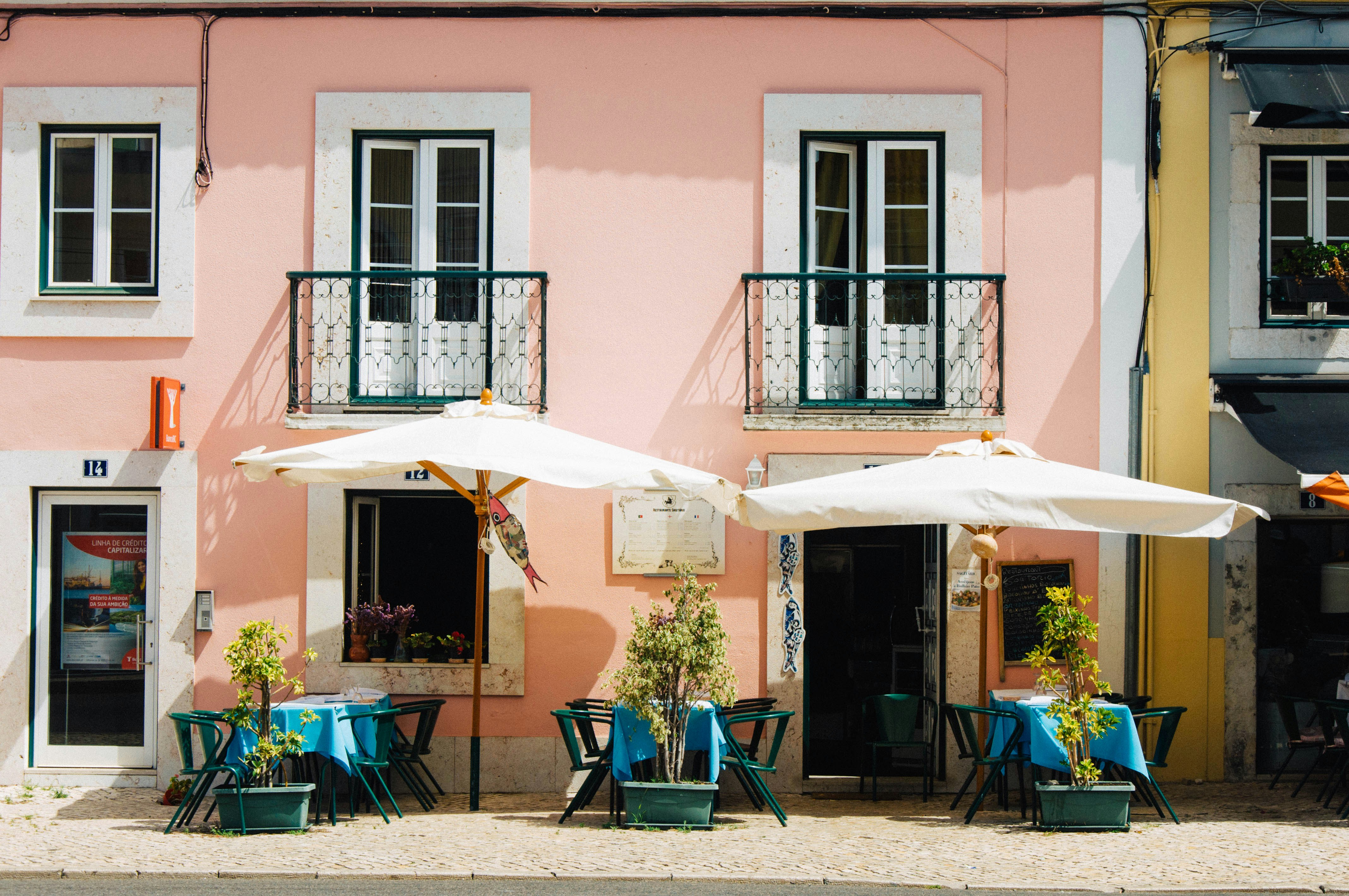 A pink building with blue tables and white umbrellas on a sidewalk patio.