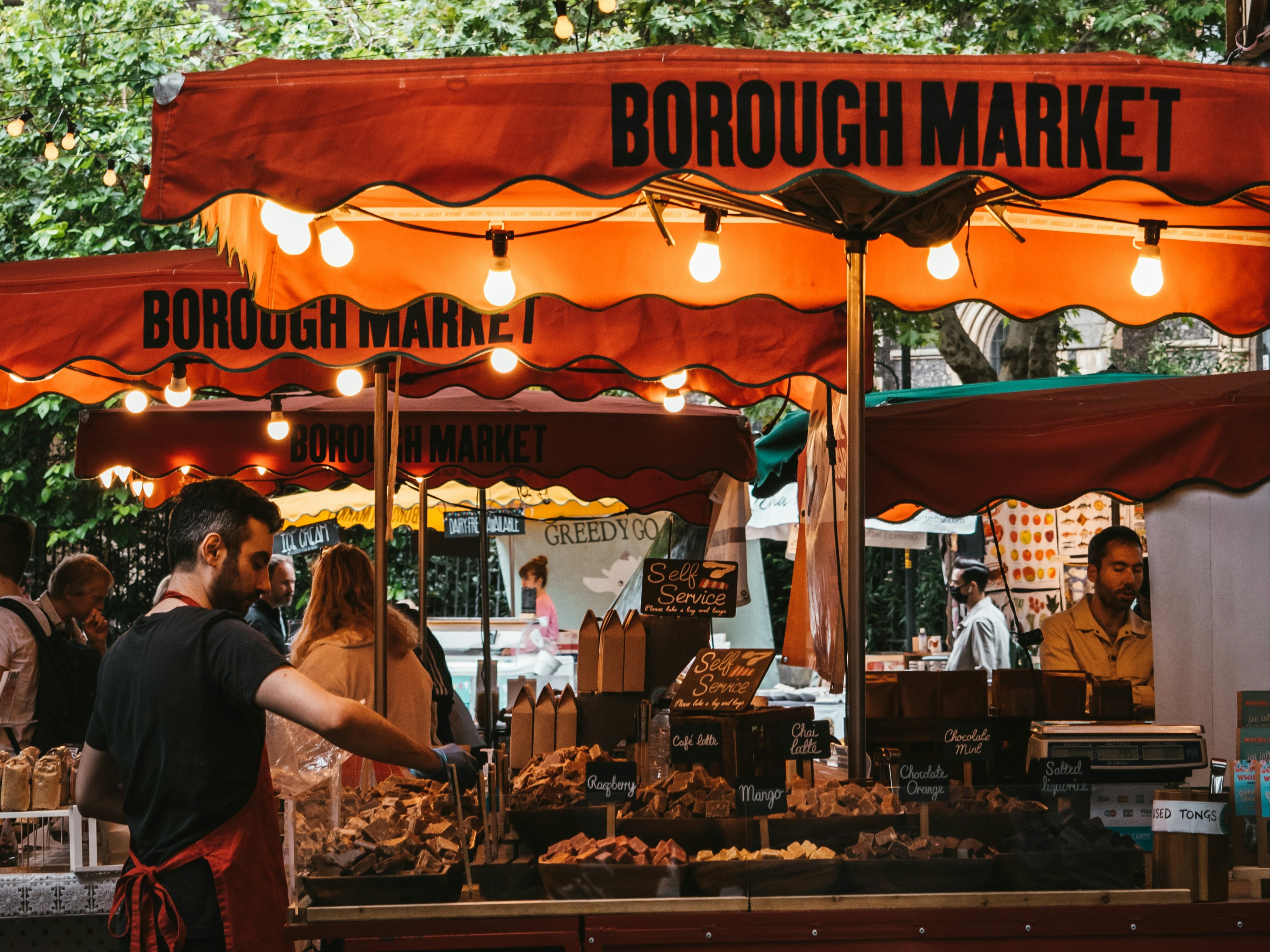 The image captures the lively atmosphere of Borough Market, with vibrant orange canopies over stalls displaying an array of breads, under strings of light bulbs.