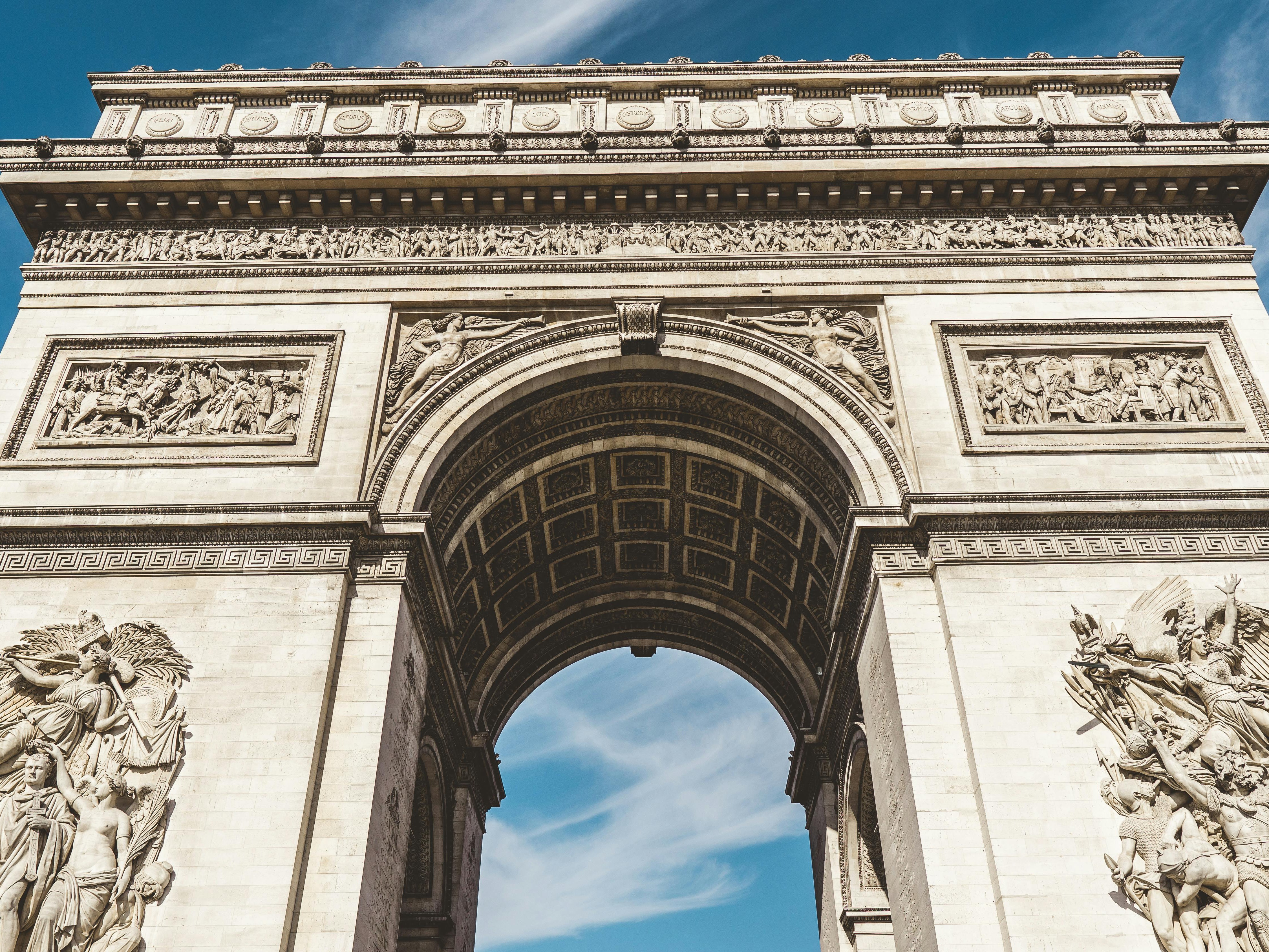 An upwards shot of the Arc de Triomphe in Paris, showcasing its detailed sculptures under a clear blue sky.
