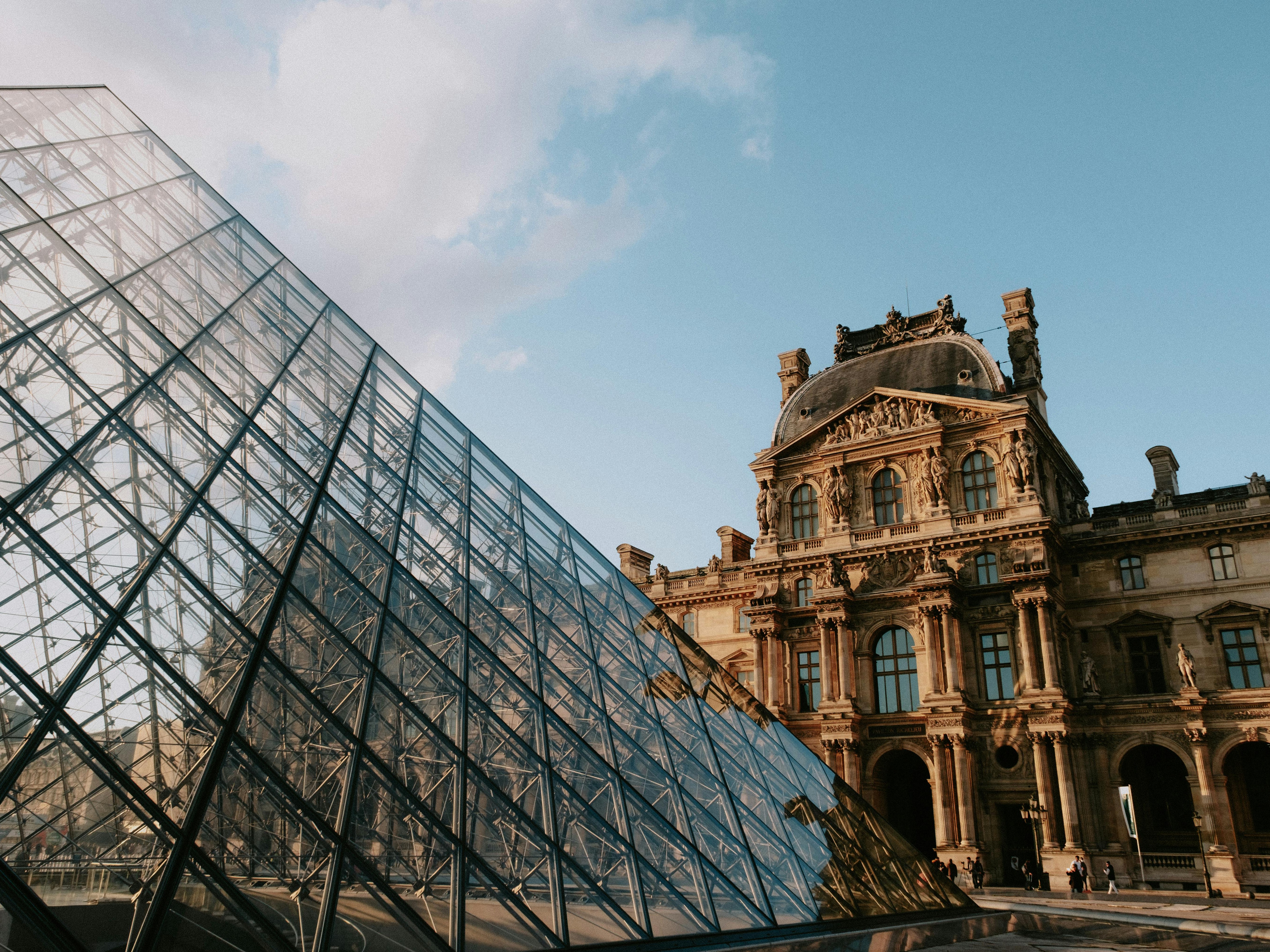 The iconic Louvre Pyramid with a glimpse of the Louvre Museum in the background under a clear sky.