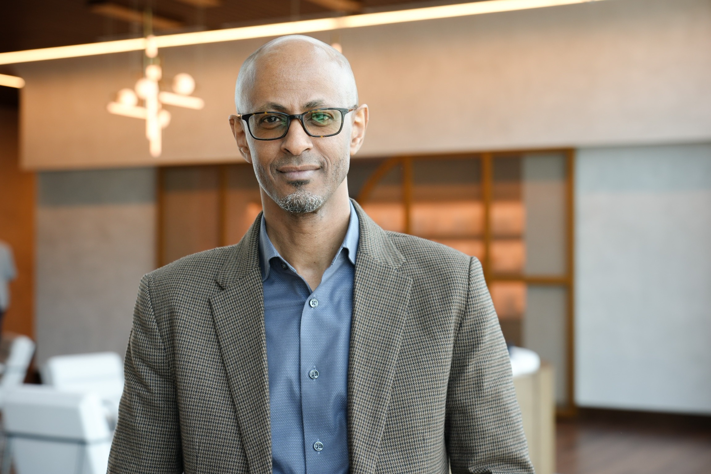 man in suit jacket and glasses standing in a hotel lobby looking at the camera