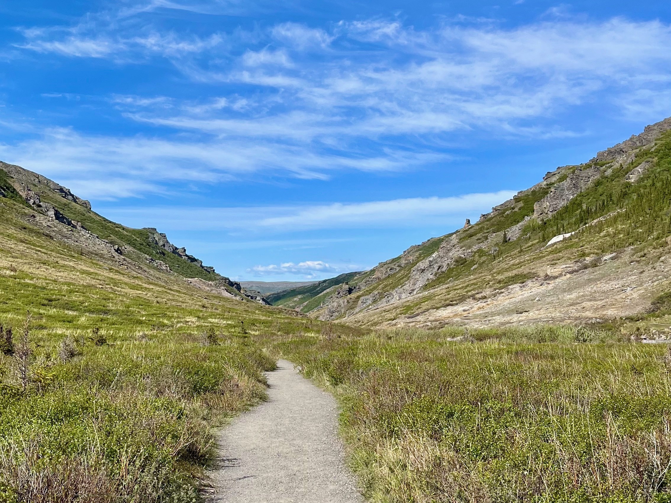 The image presents a tranquil green valley with a narrow dirt path, flanked by rocky hills under a blue sky with scattered clouds.