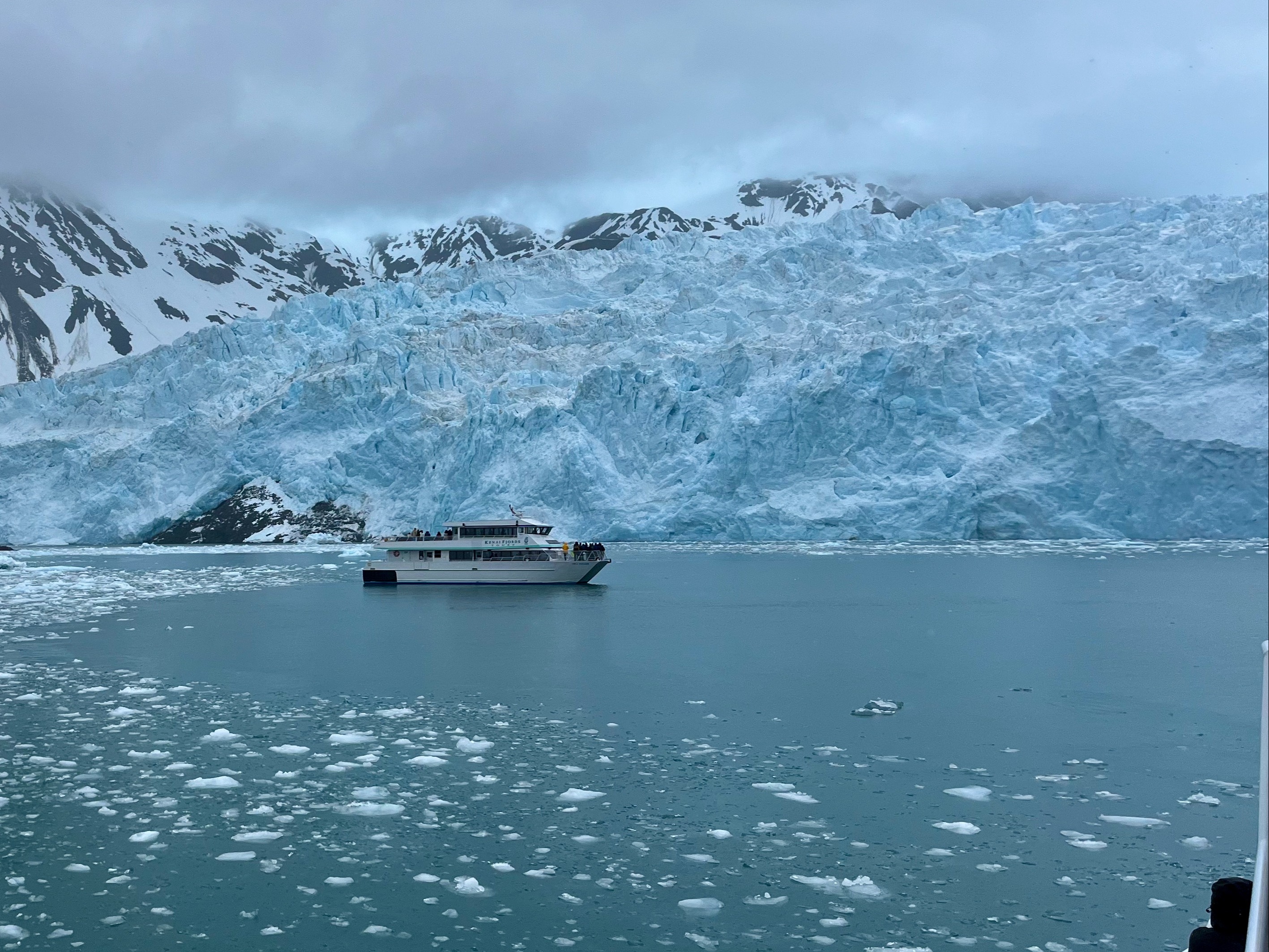 The image features a majestic glacier with deep blue hues, dwarfing a tour boat, under overcast skies and mountainous terrain.