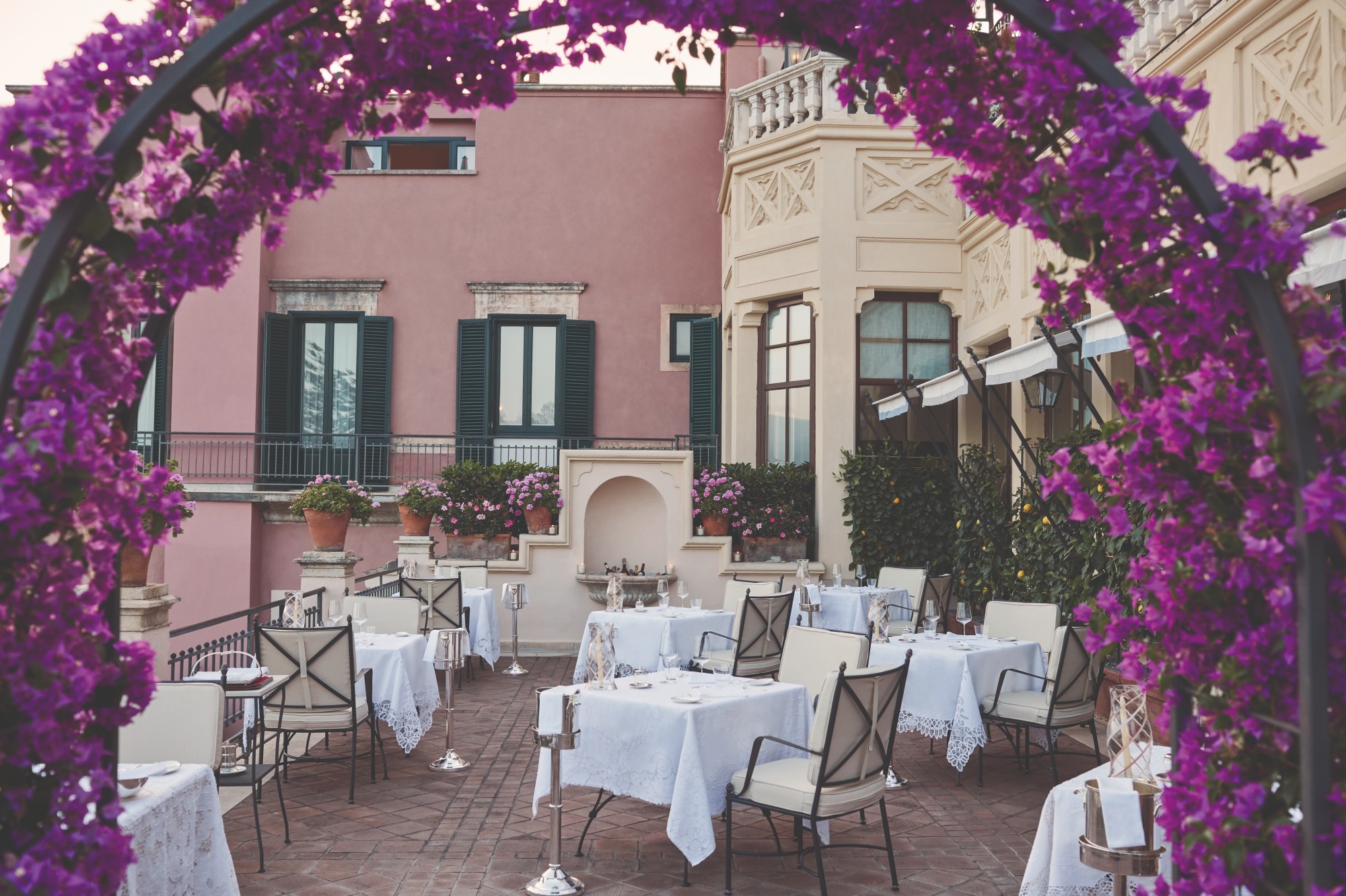 terrace with dining tables with an archway of purple flowers and a pink hotel in the background