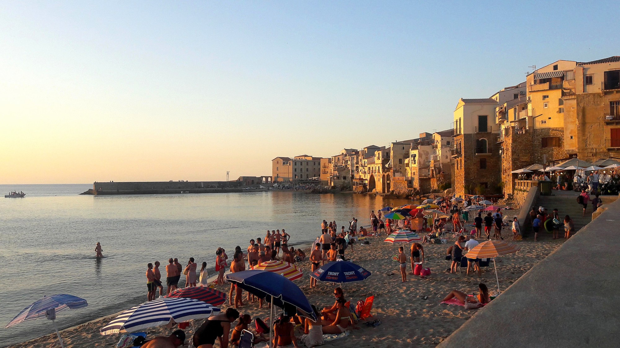 Beachgoers on the sand in Cefalù under umbrellas at sunset, with yellow buildings in the background
