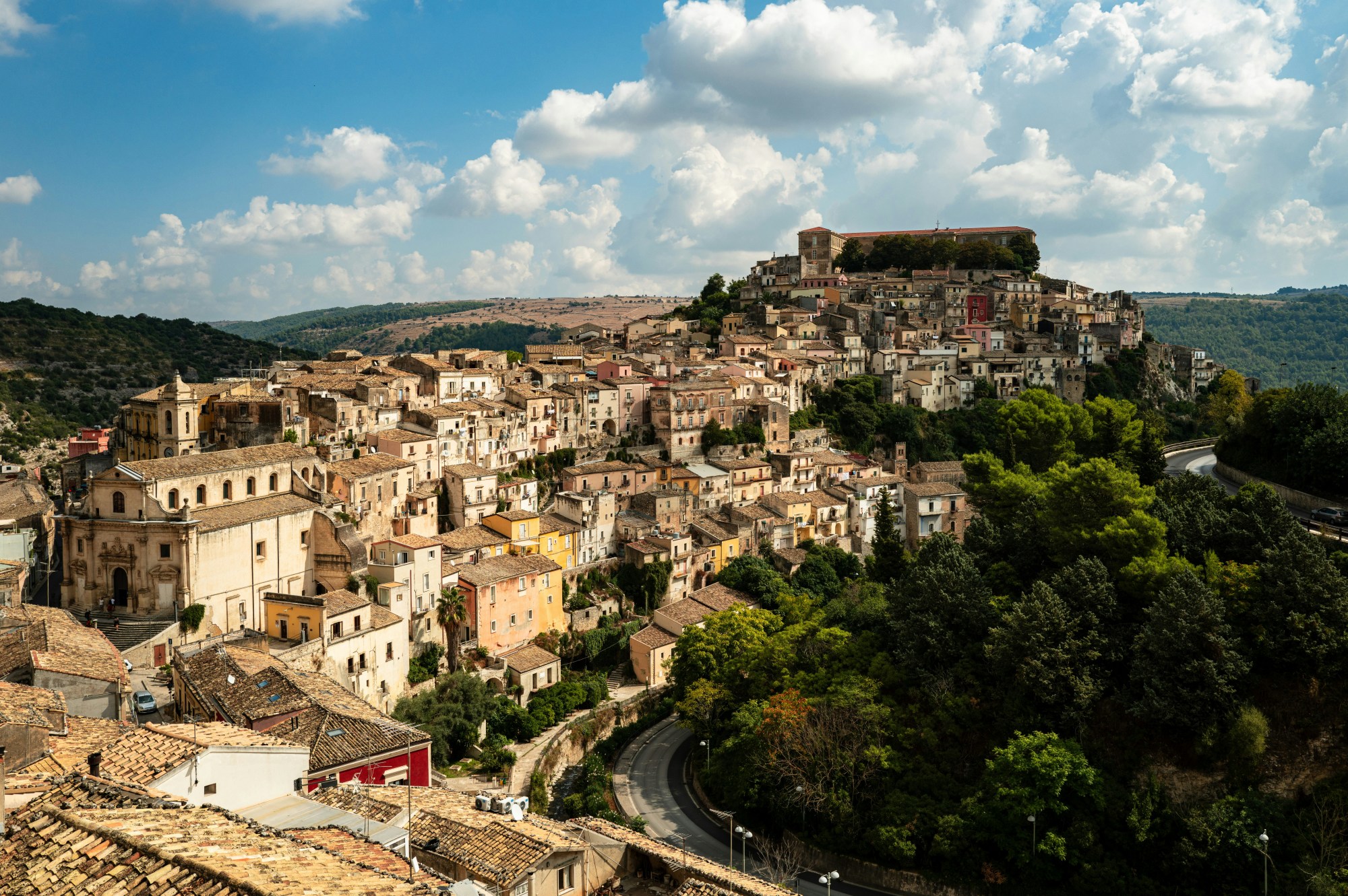 Buildings on a hillside next to a winding road with a forest on one side.
