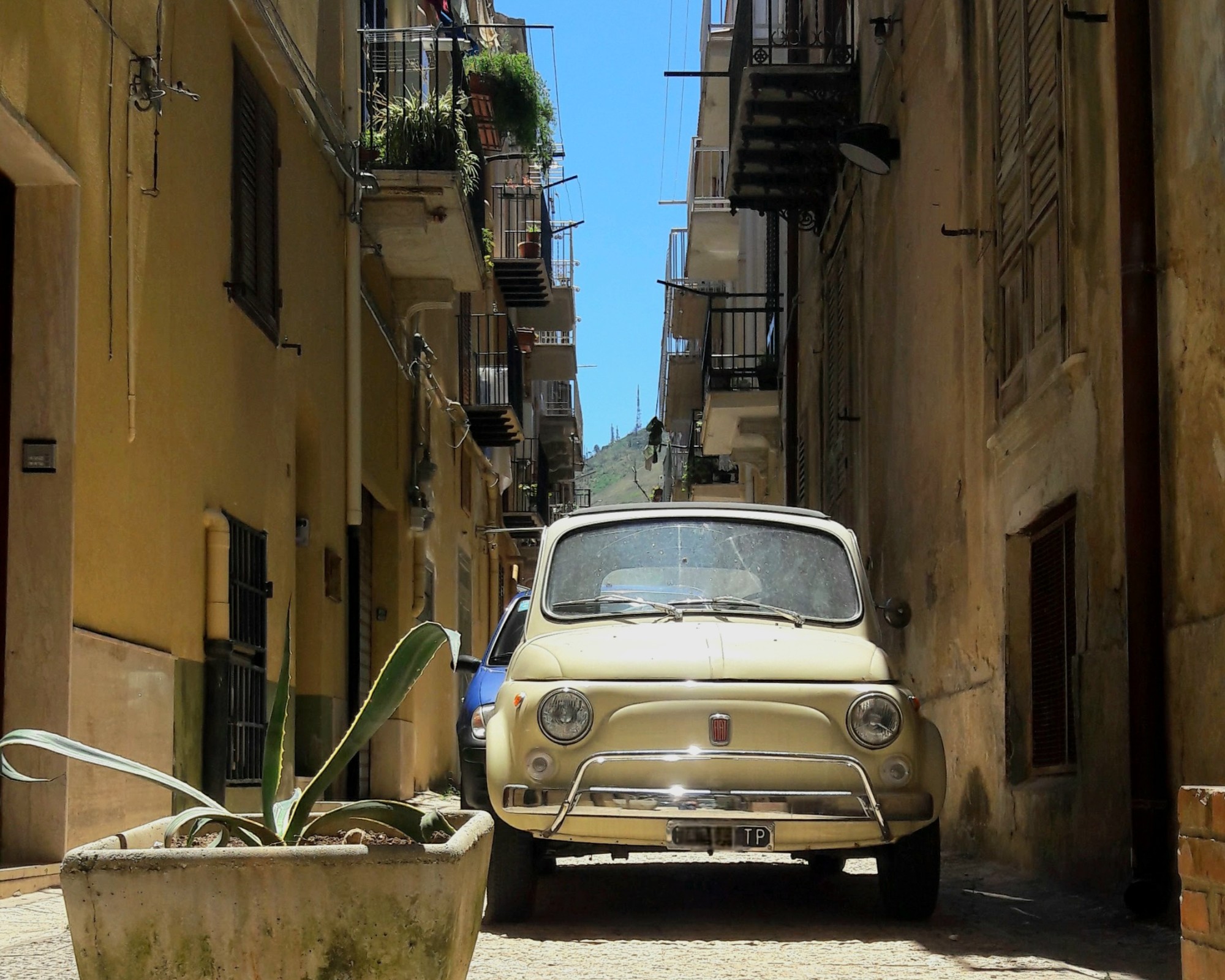 A vintage yellow car in a Sicilian alleyway behind a planter box.