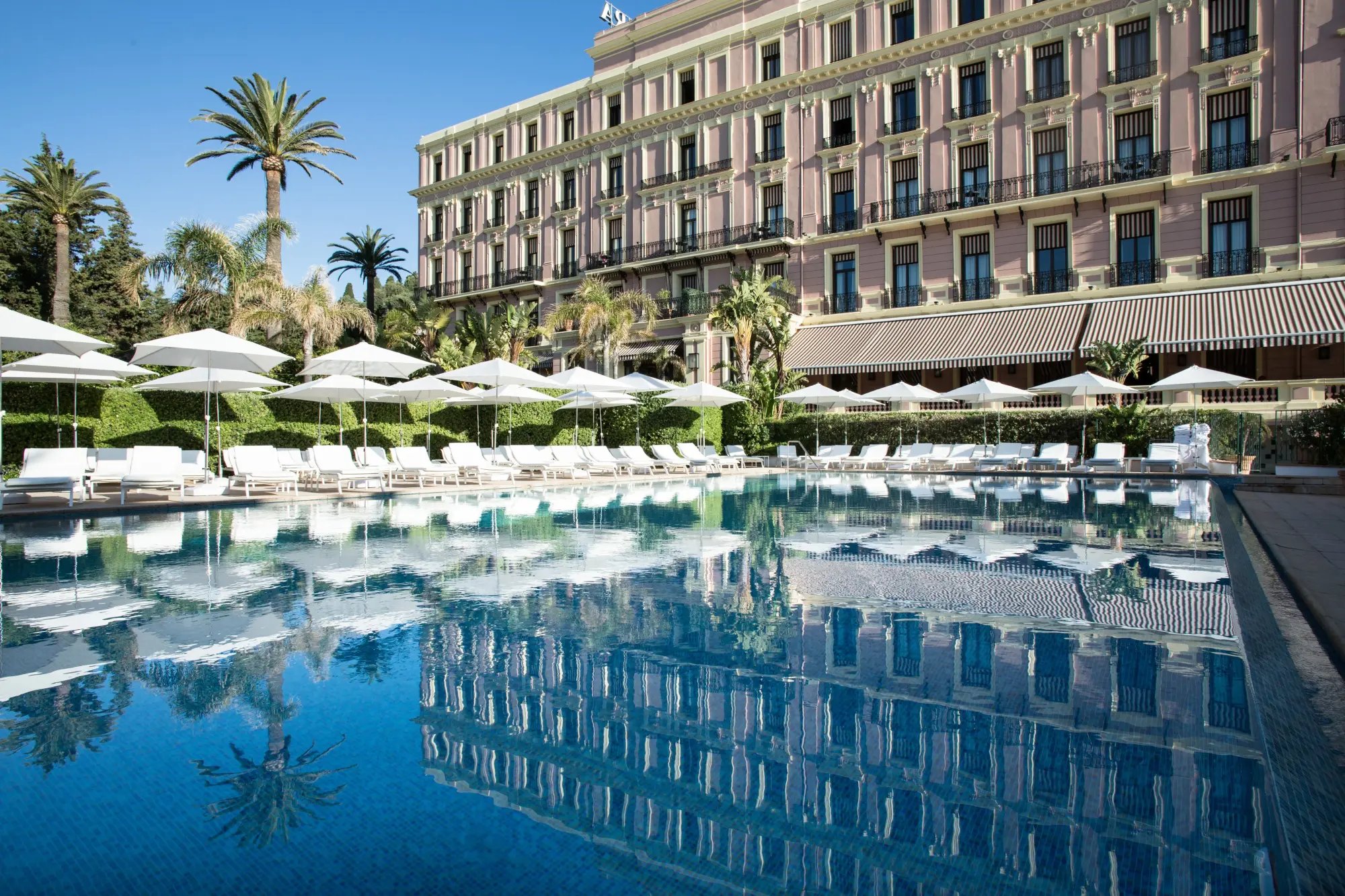 a swimming pool surrounded by white umbrellas in front of a fancy pink hotel