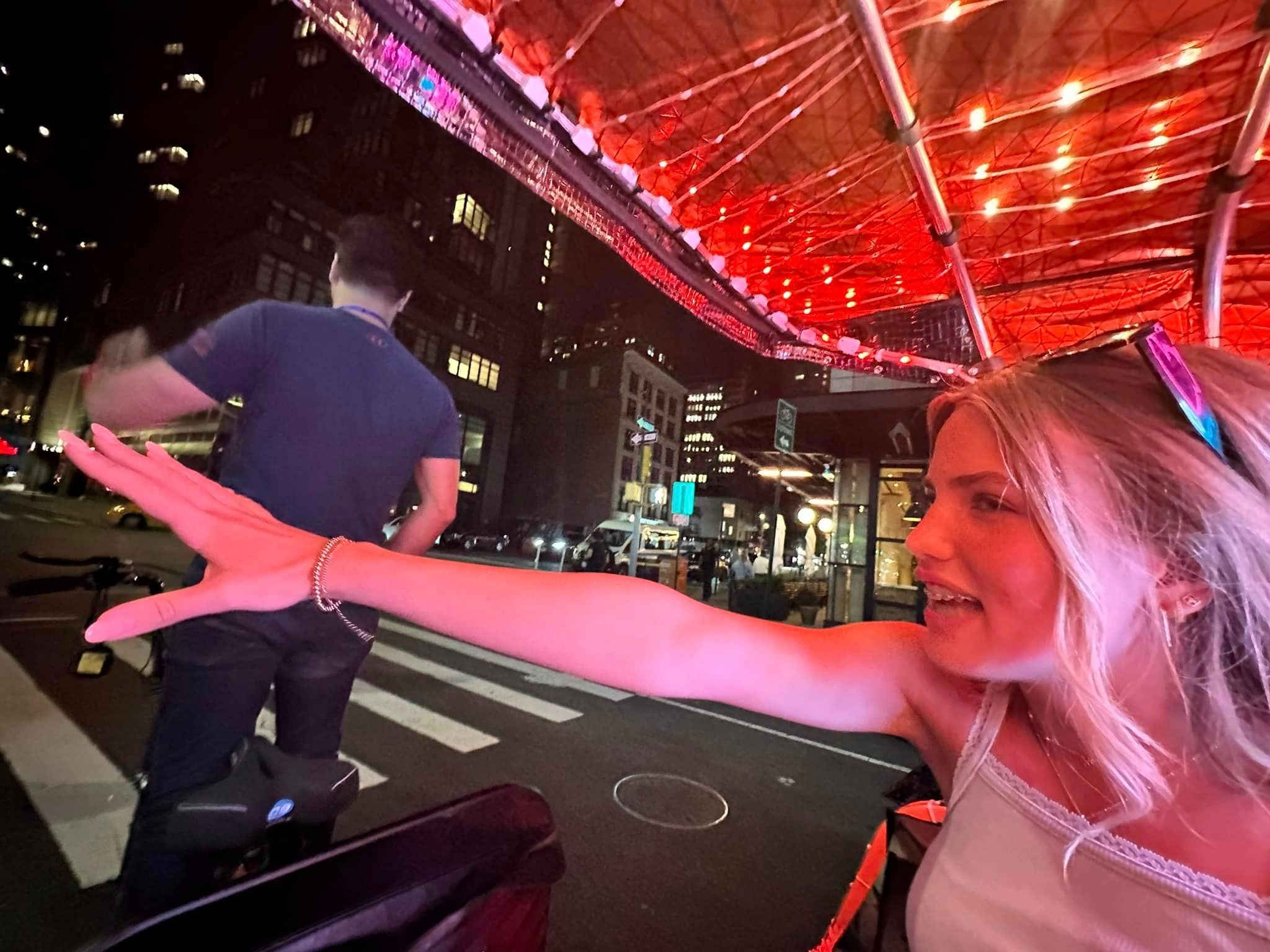 Someone sits underneath an awning with red lights at nighttime with their arm extended out towards the sidewalk.