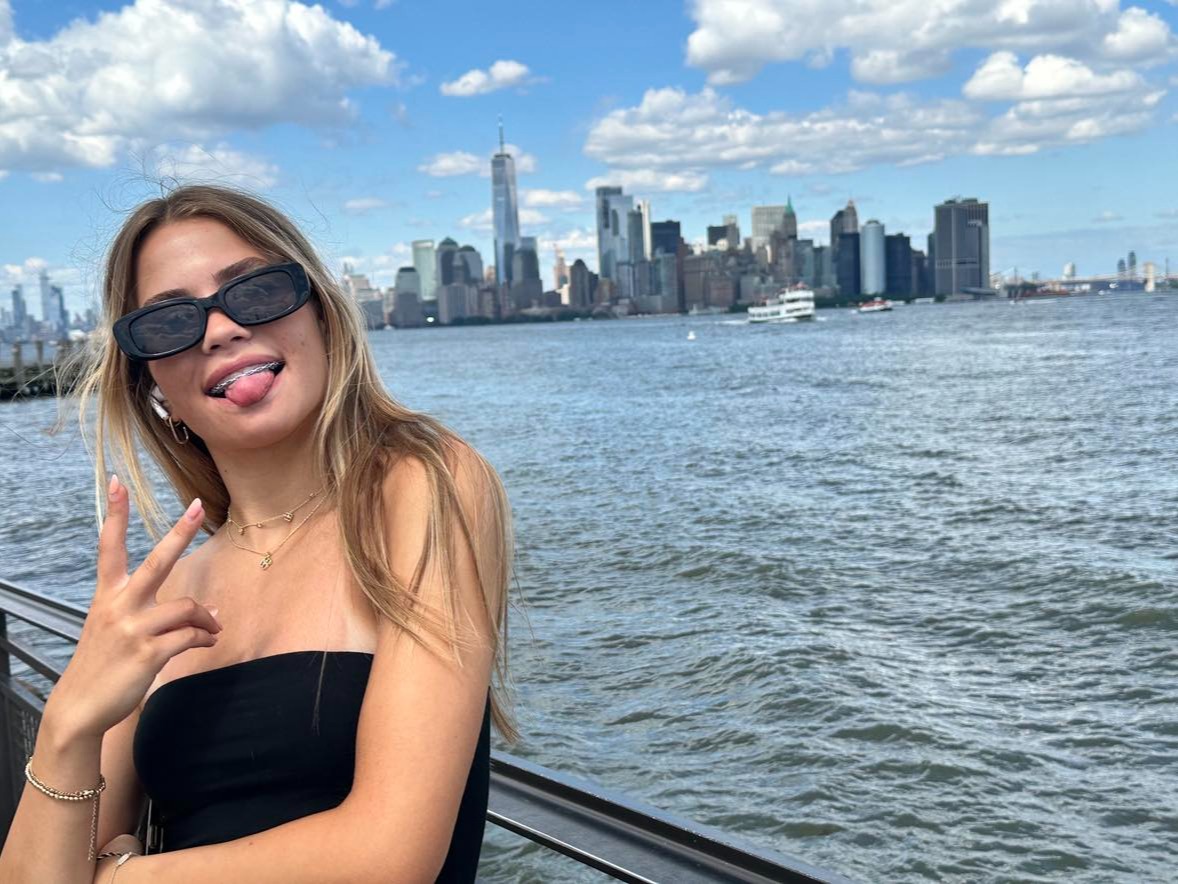A person posing in front of the river with her tongue out and sunglasses on with the New York skyline in the background.