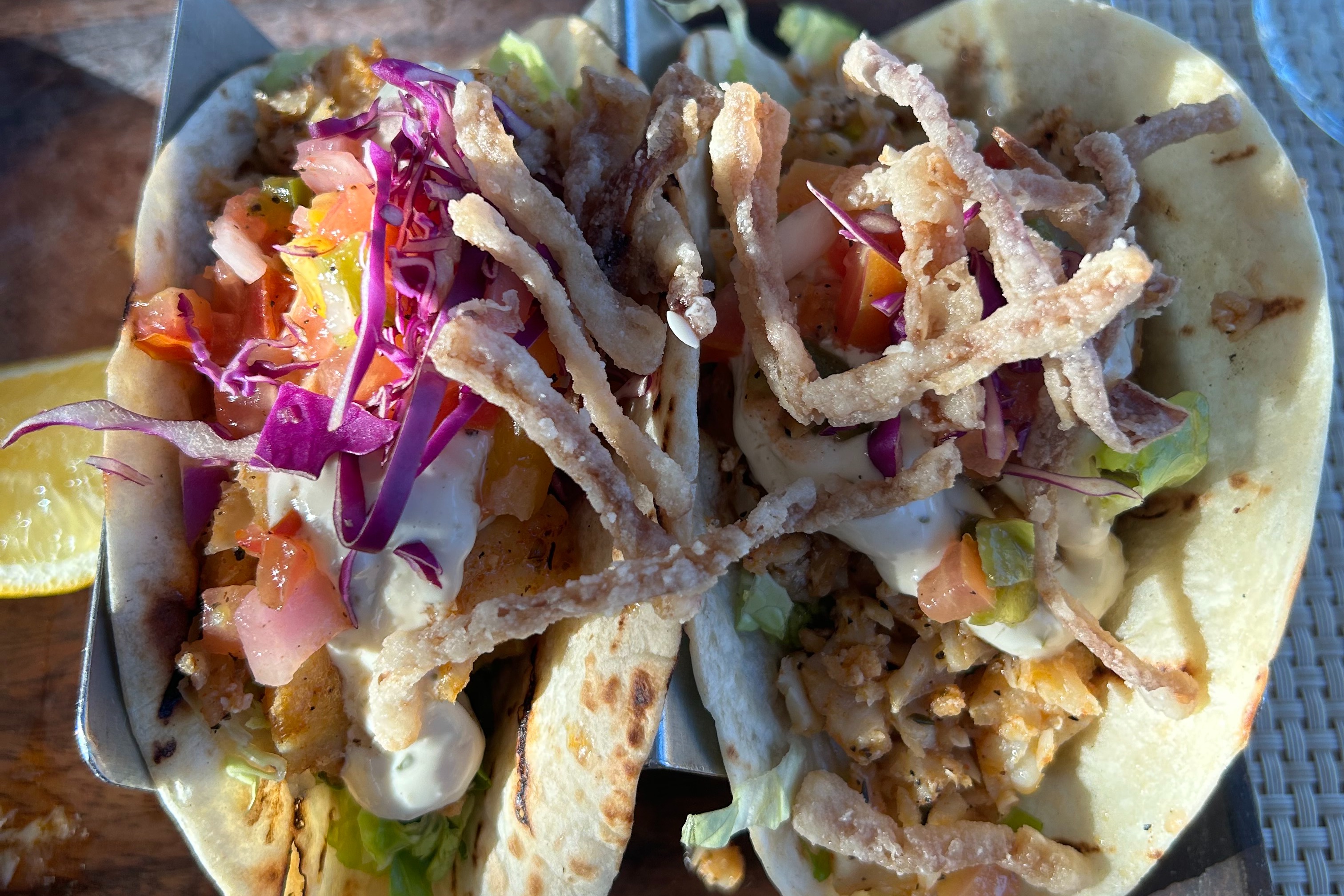 Two fish tacos standing in a metal taco holder and garnished with different colorful veggies.