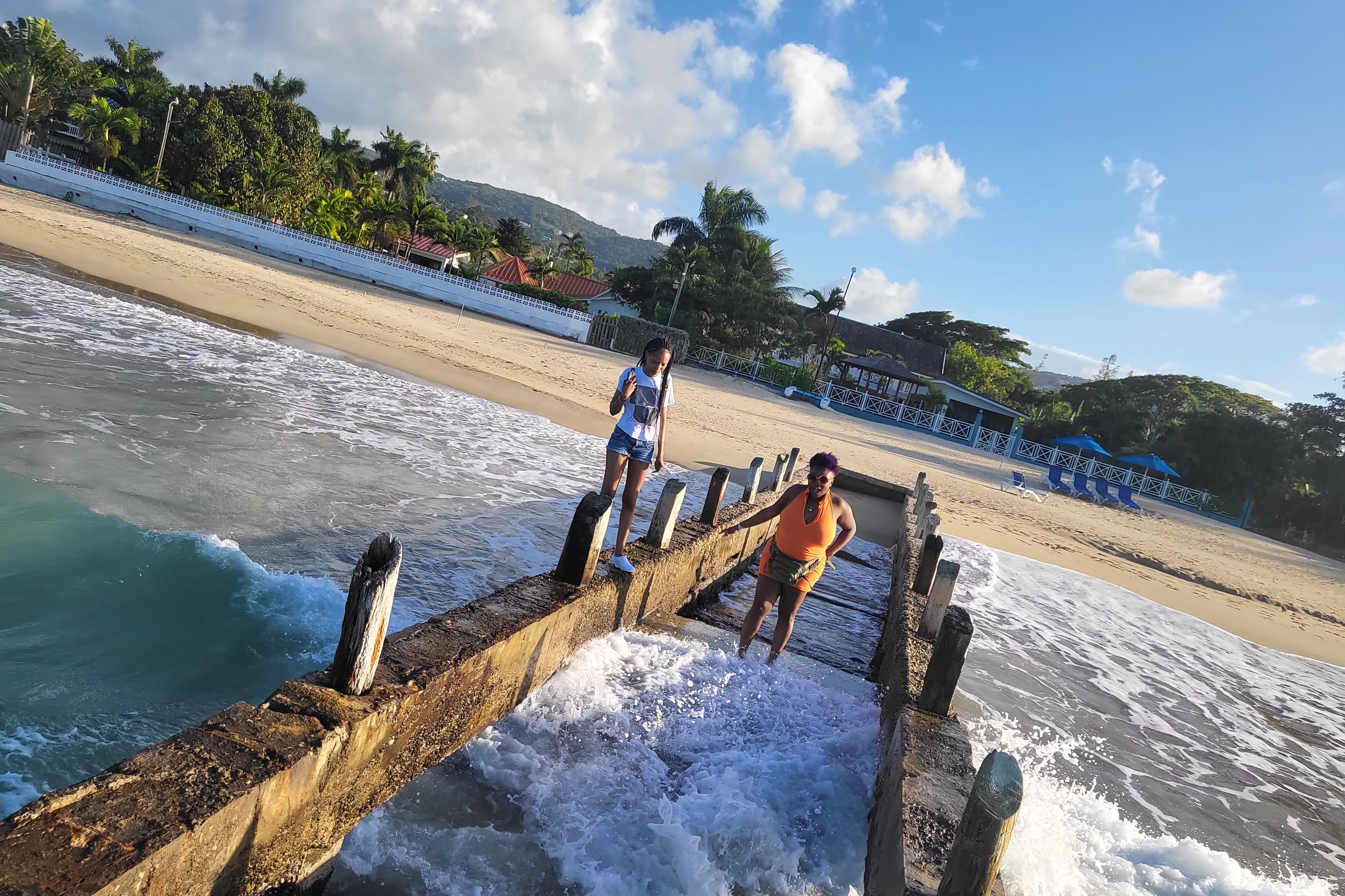 Two people enjoying waves splashing over them while entering the sea via a pathway from the shore.