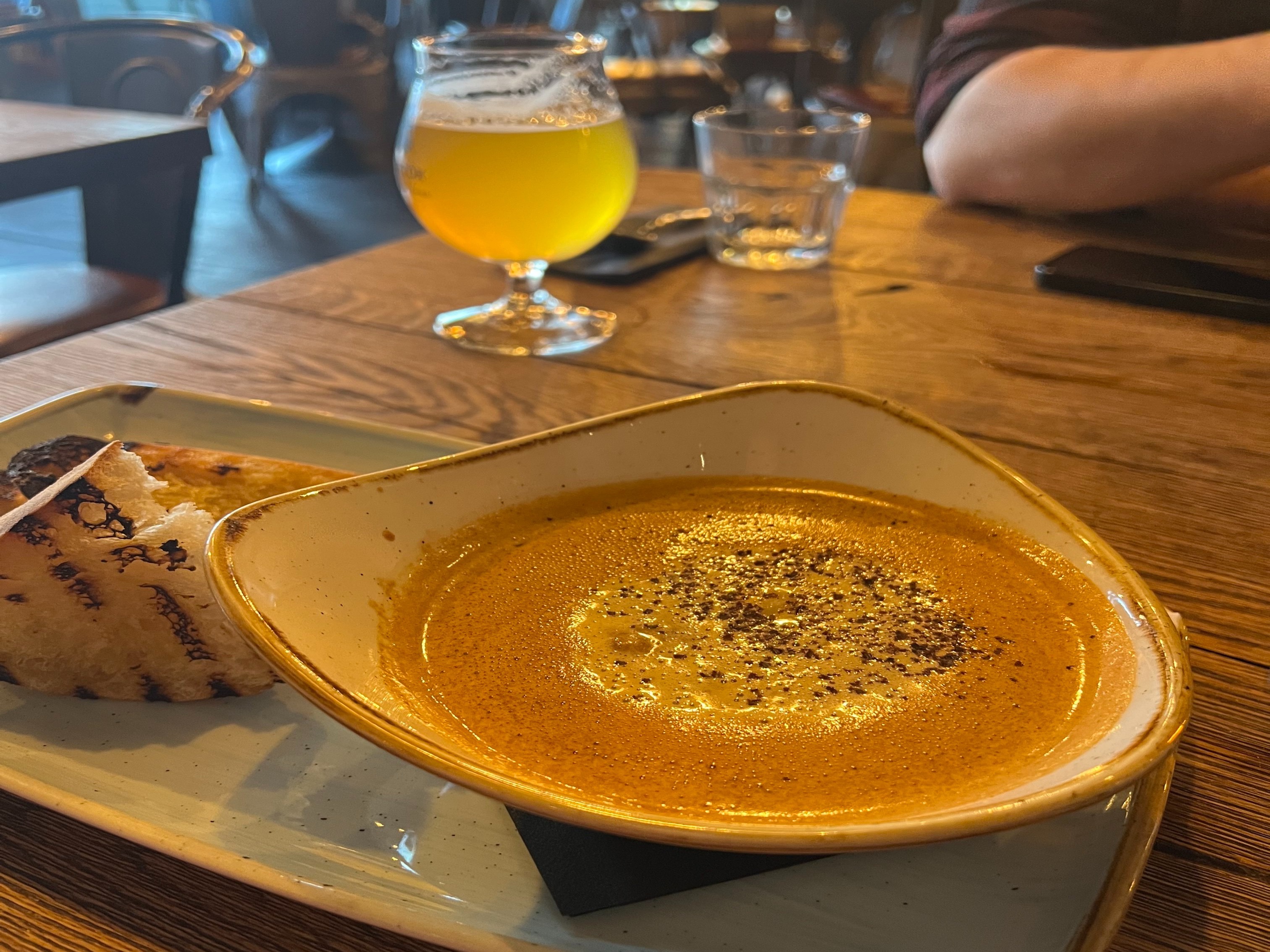 A bowl of soup with bread and a glass of beer served on a wooden table at a restaurant