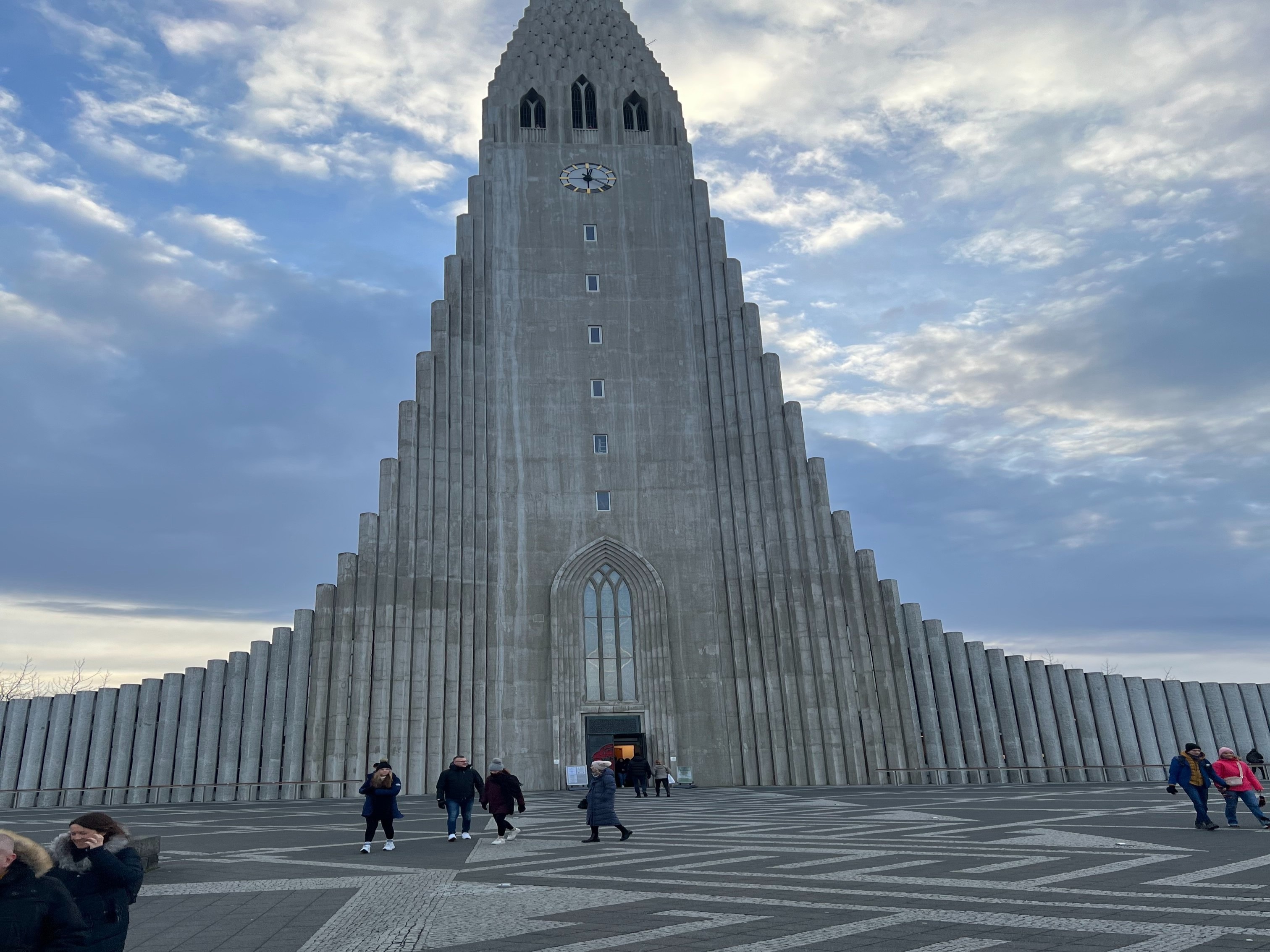 A grand church with a stepped concrete facade under a cloudy sky, with people walking in front