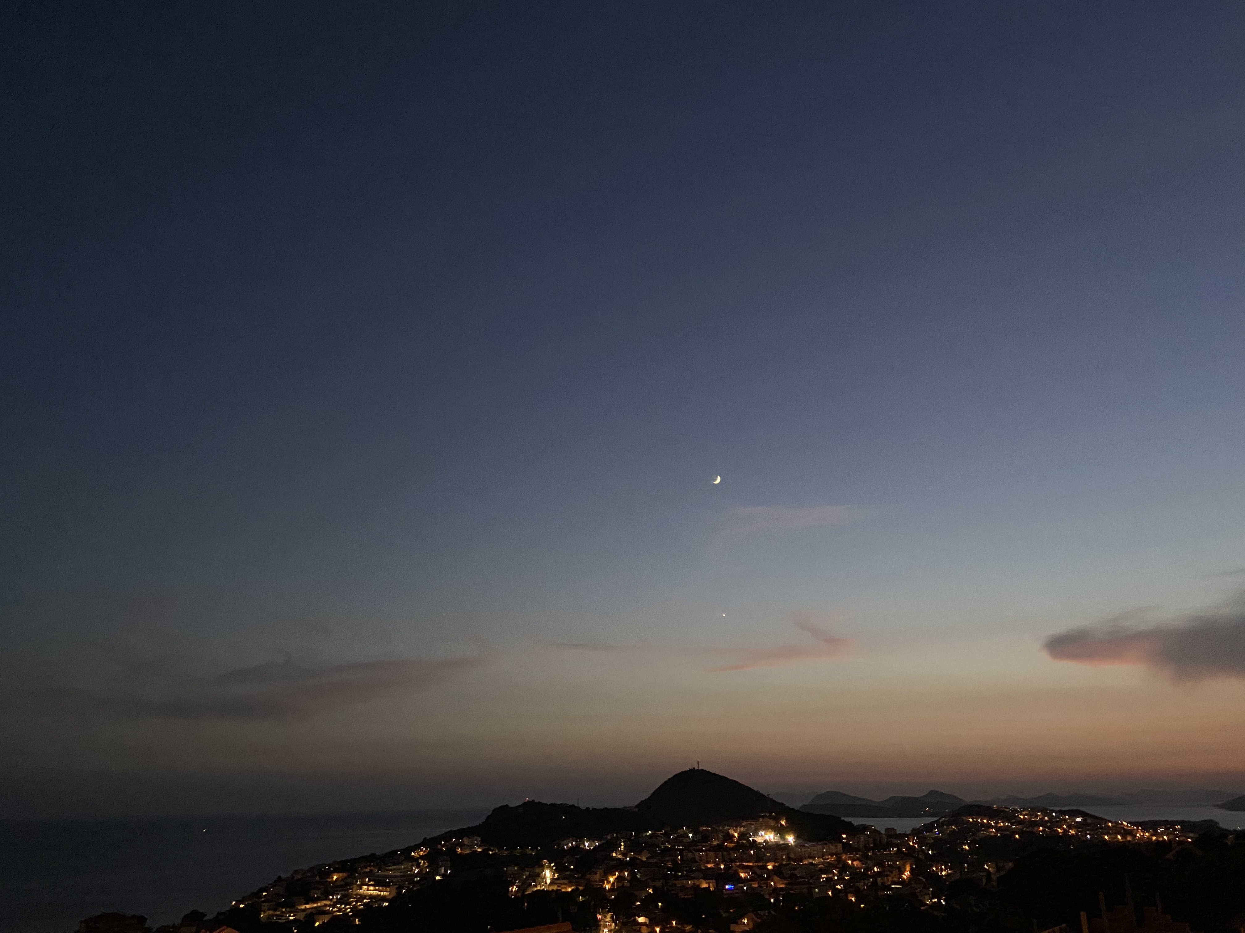 A tranquil twilight scene with a crescent moon hovering over a silhouetted landscape and a lit-up town.