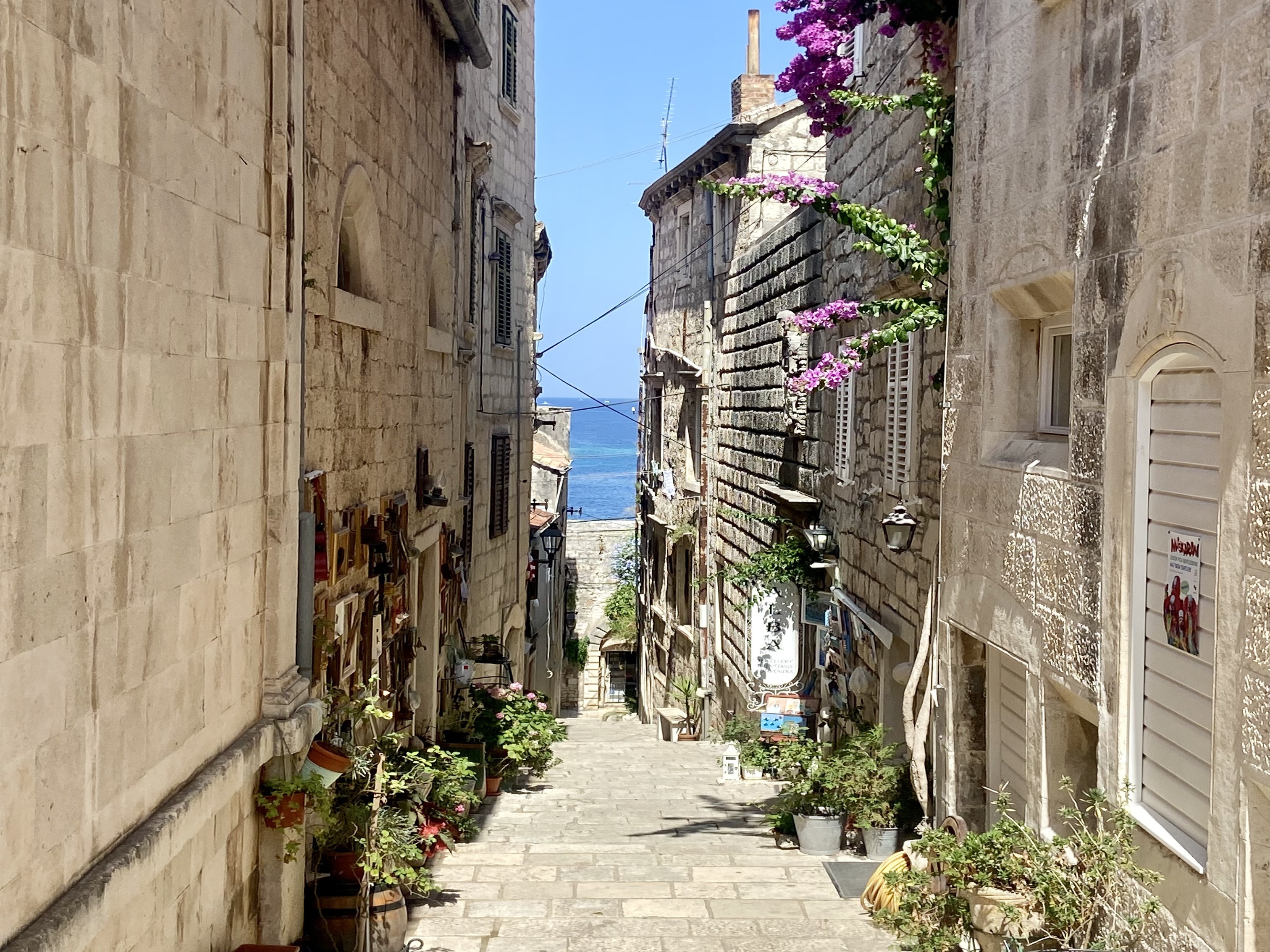 A charming cobblestone street lined with historic stone buildings and balconies, some adorned with flowers, under a clear blue sky.