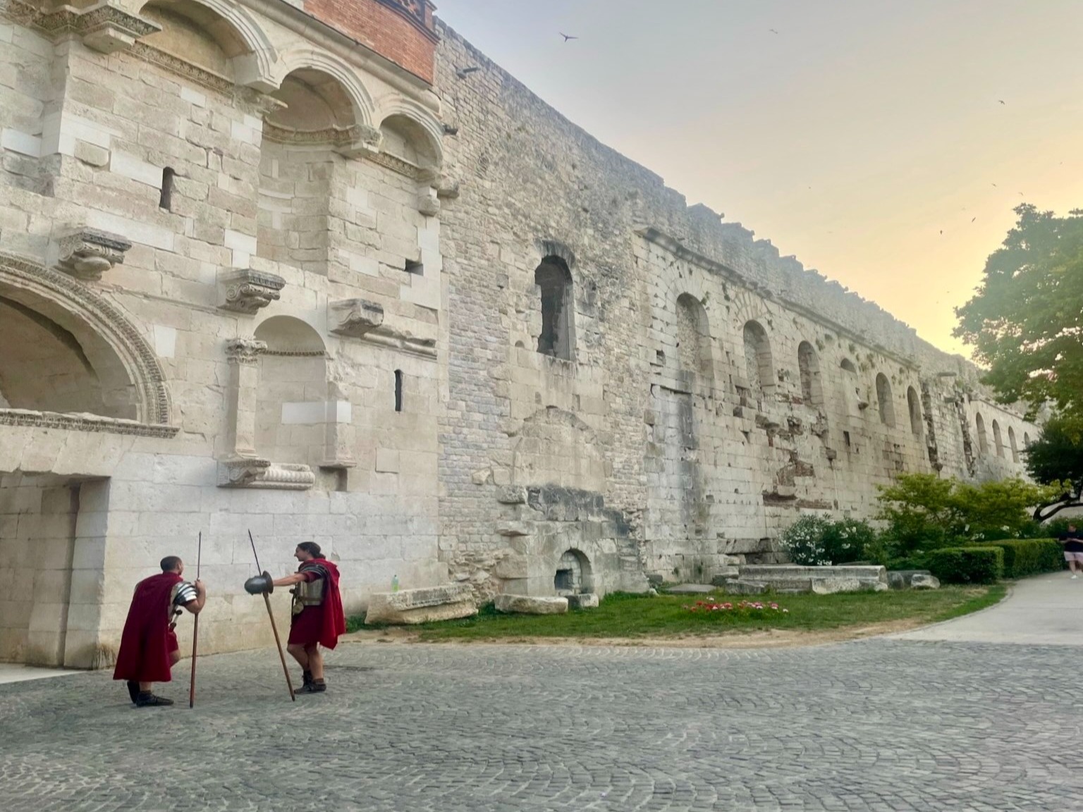 Two figures in historical attire beside an ancient wall with arches under a sunset sky,