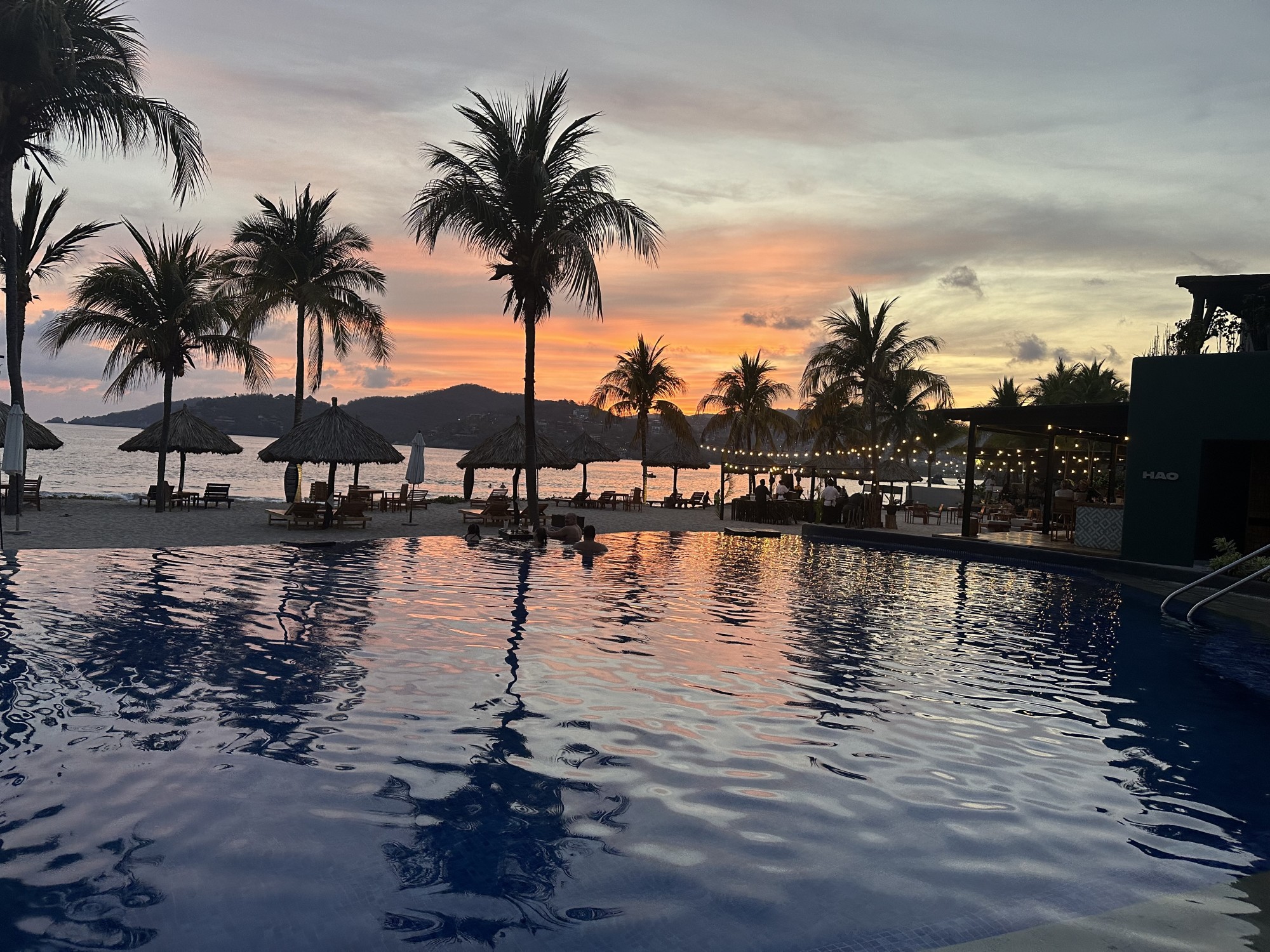 A sunset over an outside swimming pool surrounded by silhouettes of palm trees