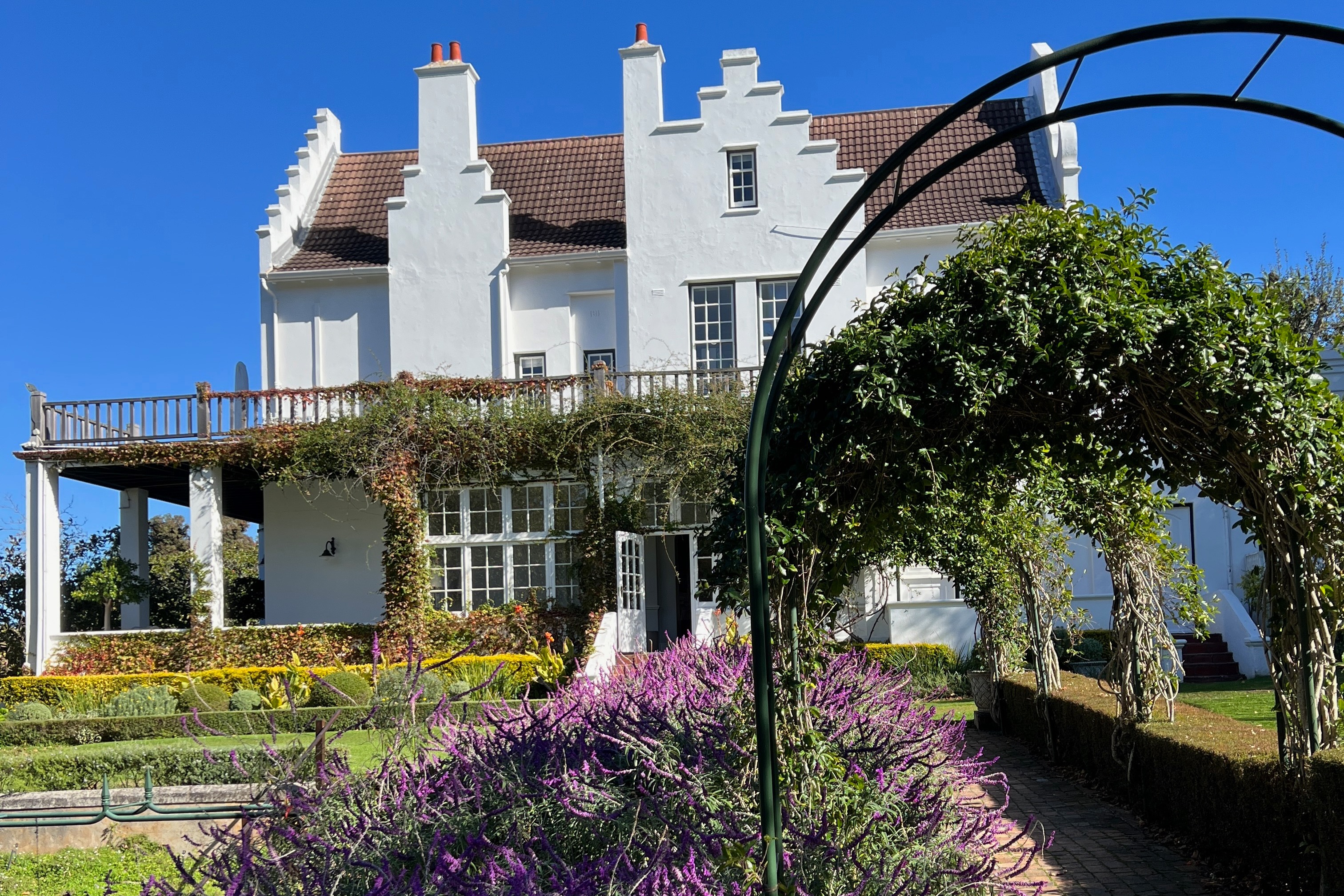 A view of a beautiful garden with trees and colorful flowers and a large white residence behind.
