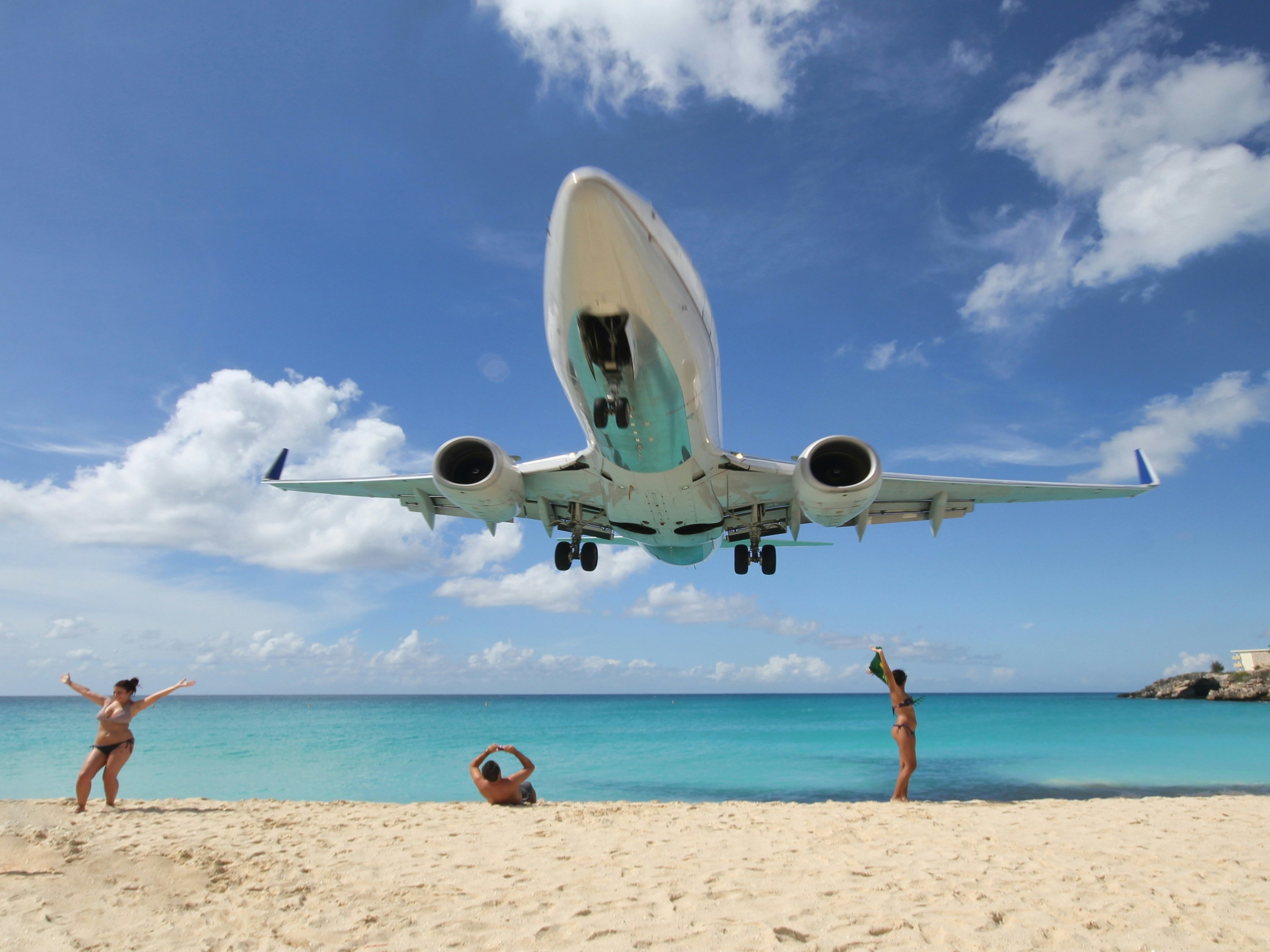 An airplane descends closely over a beautiful beach with people looking on.