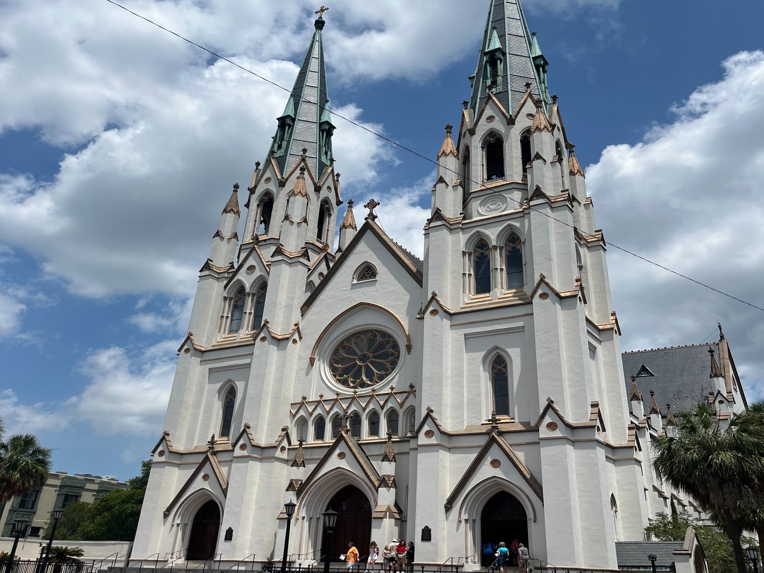 The image captures the grandeur of a large, ornate church with Gothic features against a partly cloudy sky, with people at the bottom hinting at its scale.