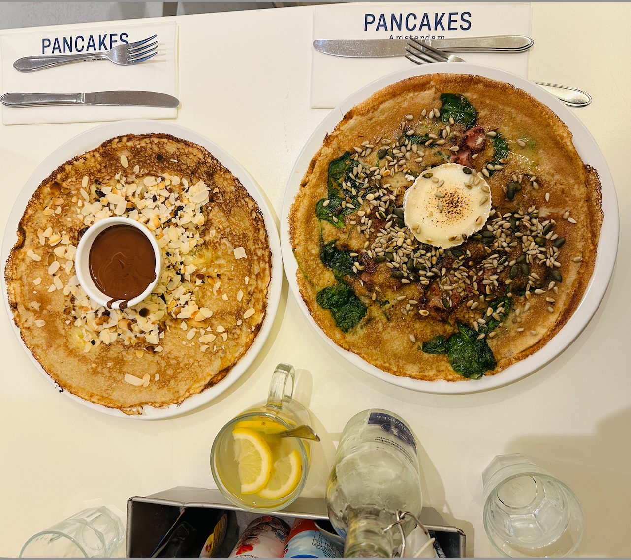 Two pancakes, one with spinach and seeds and the other with almond shavings and Nutella, on a table with cutlery.