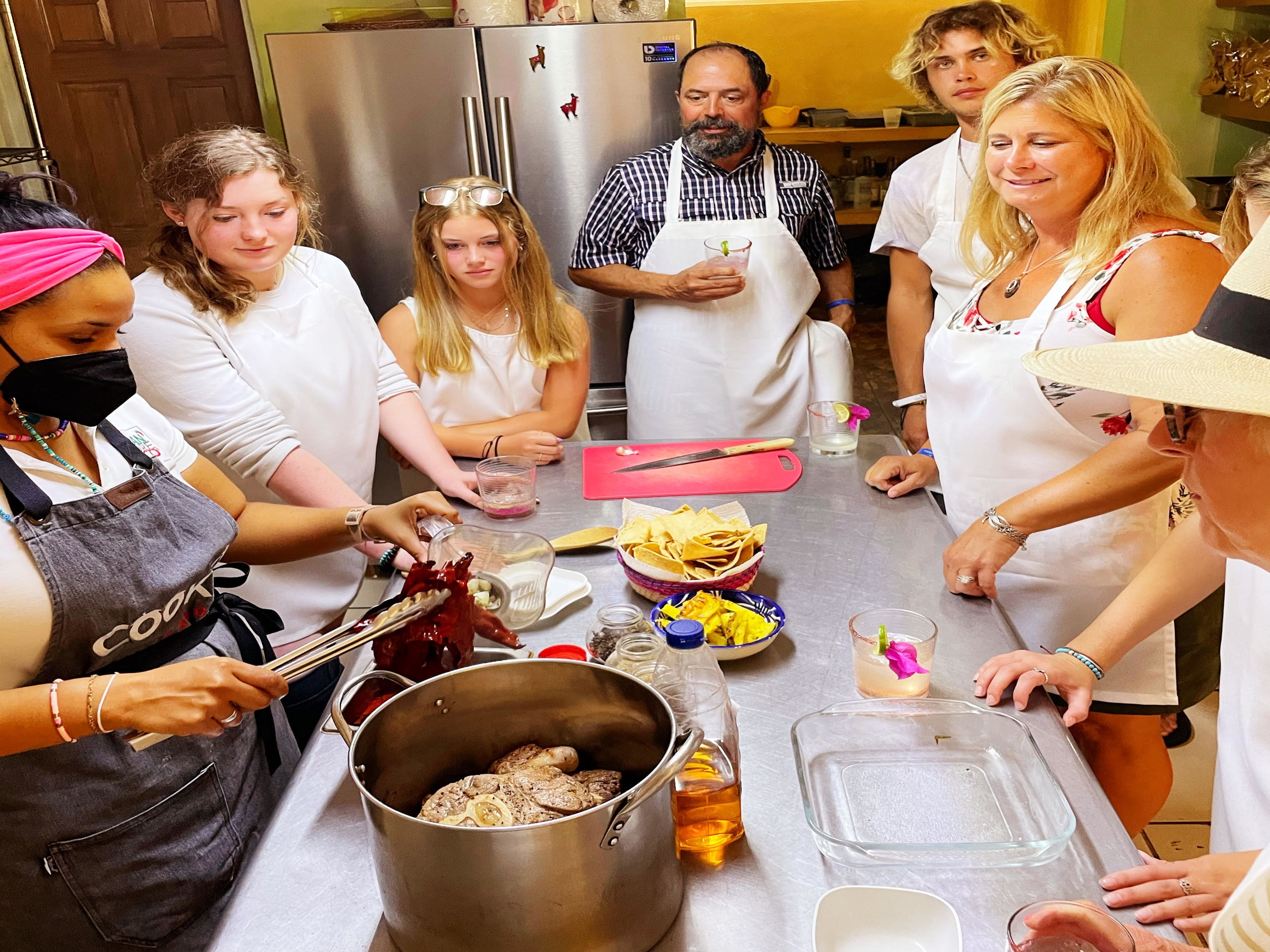 A family gathered around a metal table in a kitchen watching an instructor in an apron.