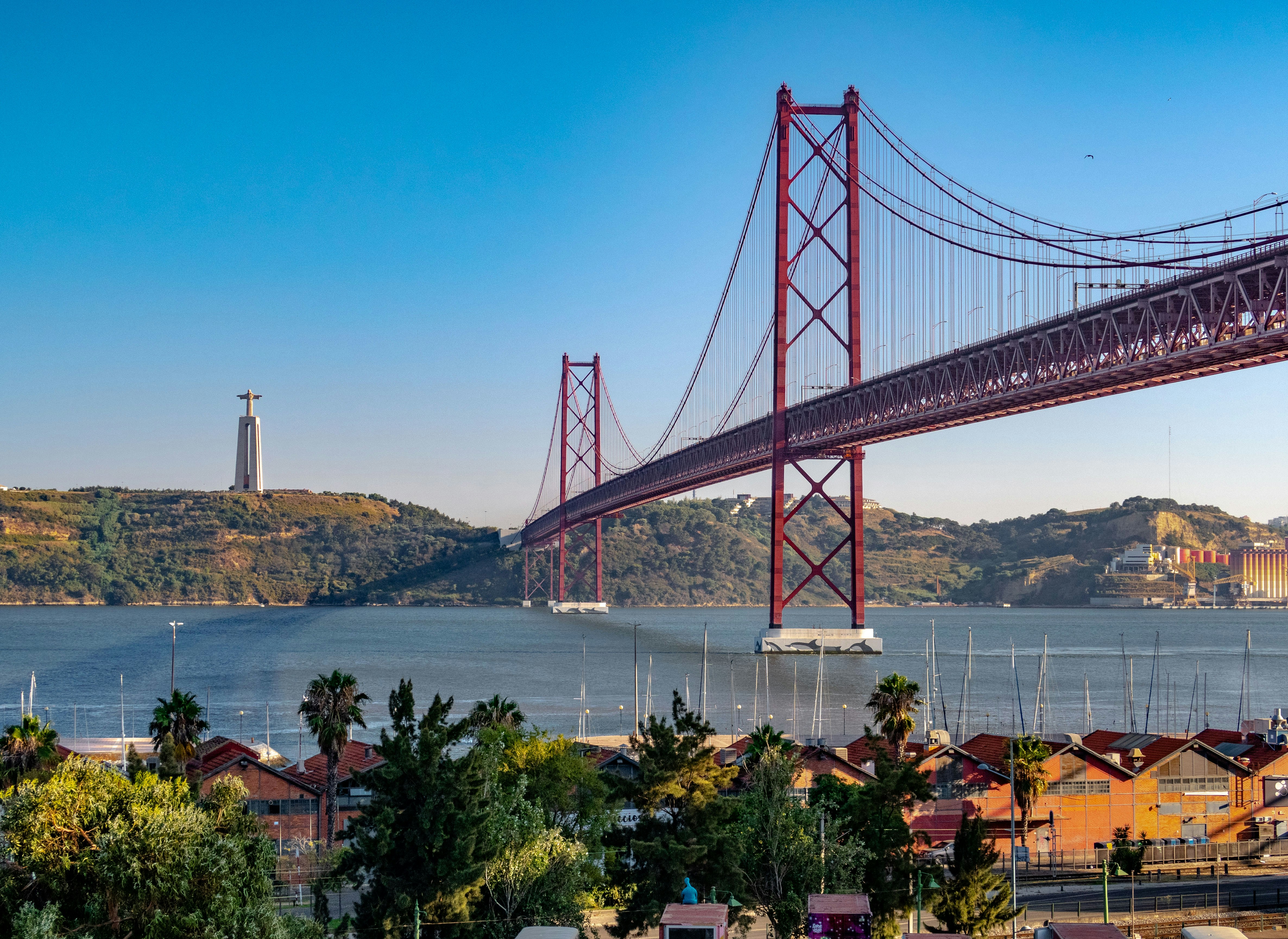 The image features a large red suspension bridge spanning over water with a statue on a hill in the background and boats docked in the foreground.