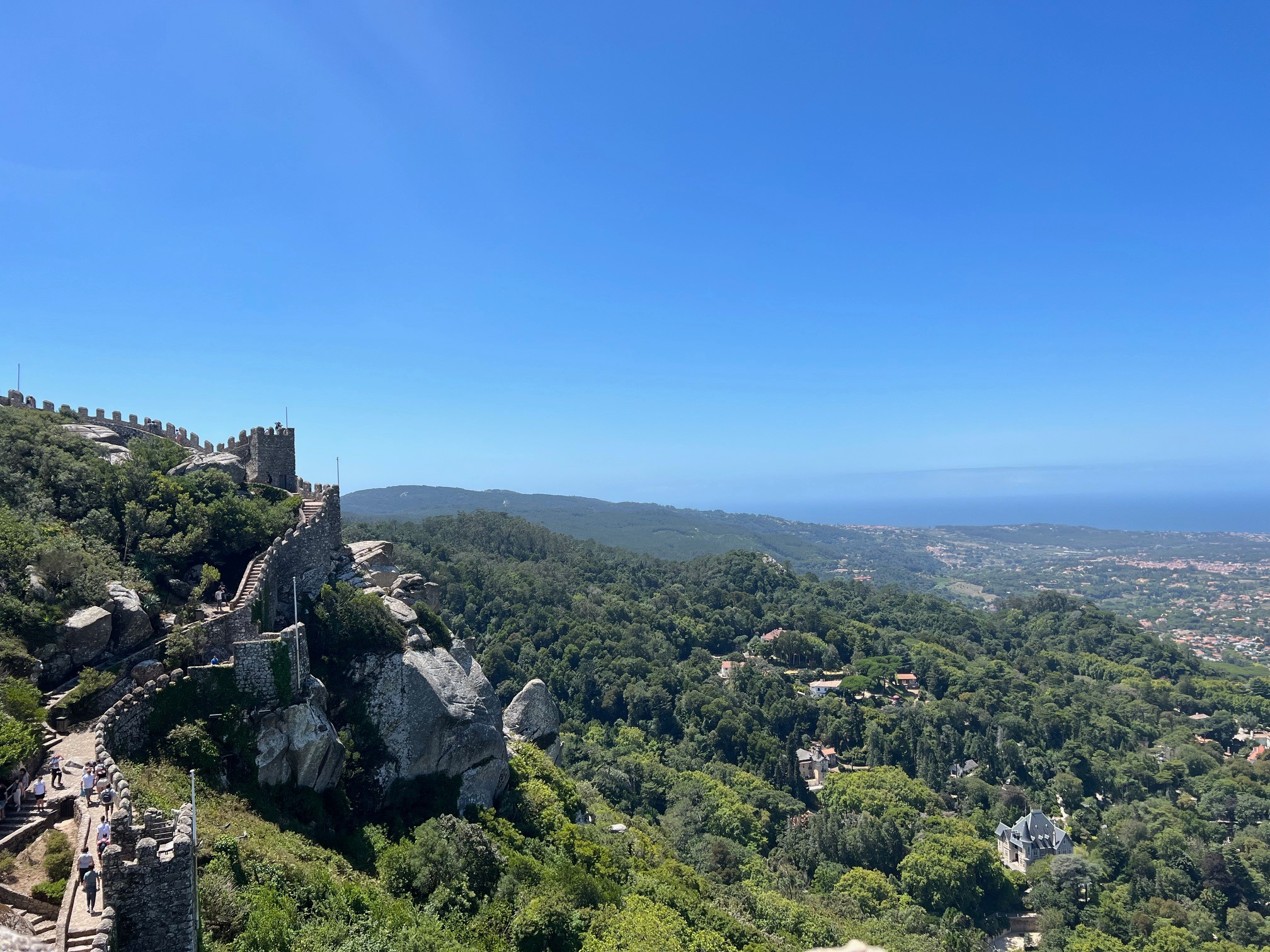 The image features a panoramic view of a castle on a hill with a stone wall extending into a lush green landscape under a clear blue sky.