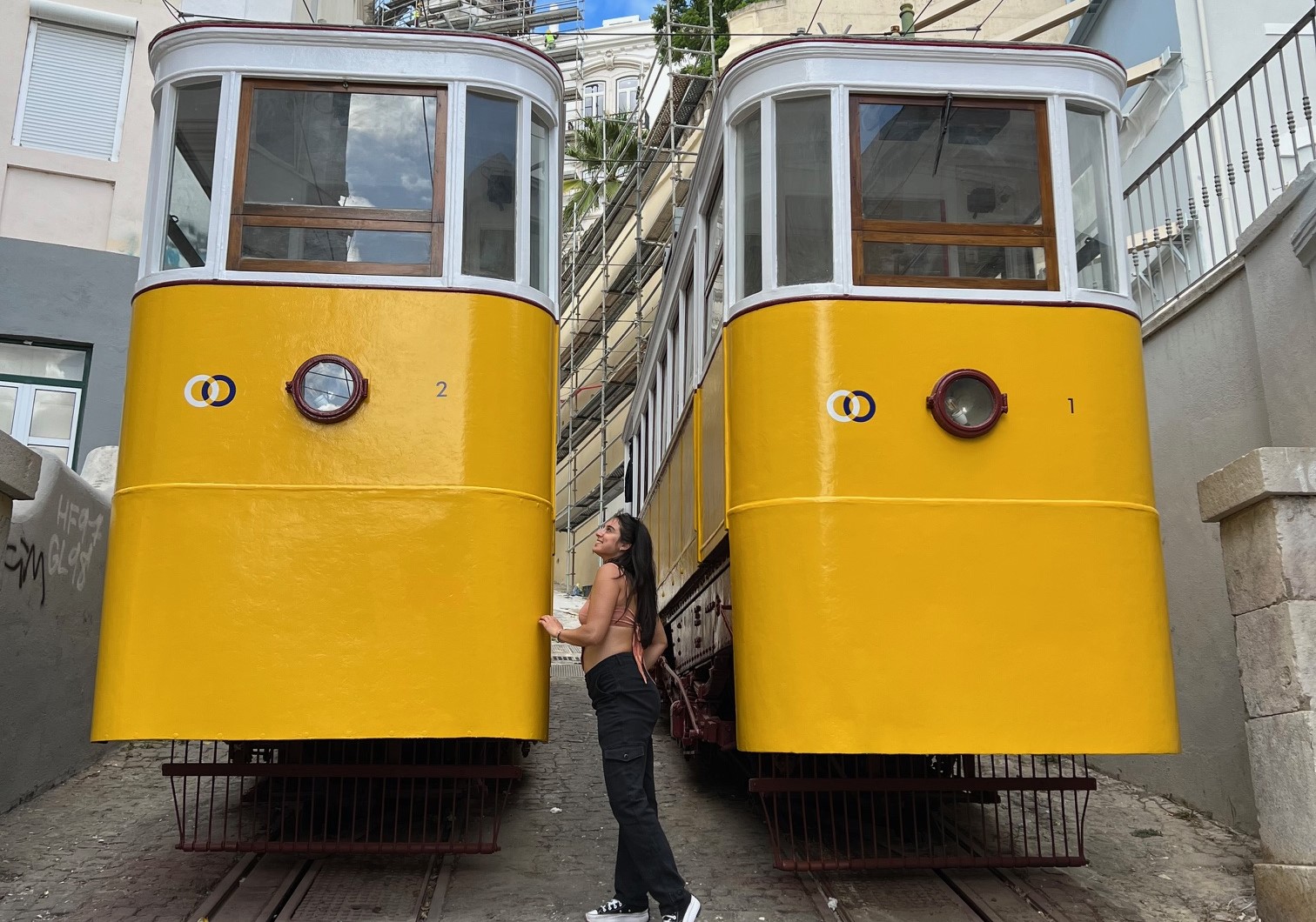 The image features two yellow trams on a street, with a person in between and buildings in the backdrop.