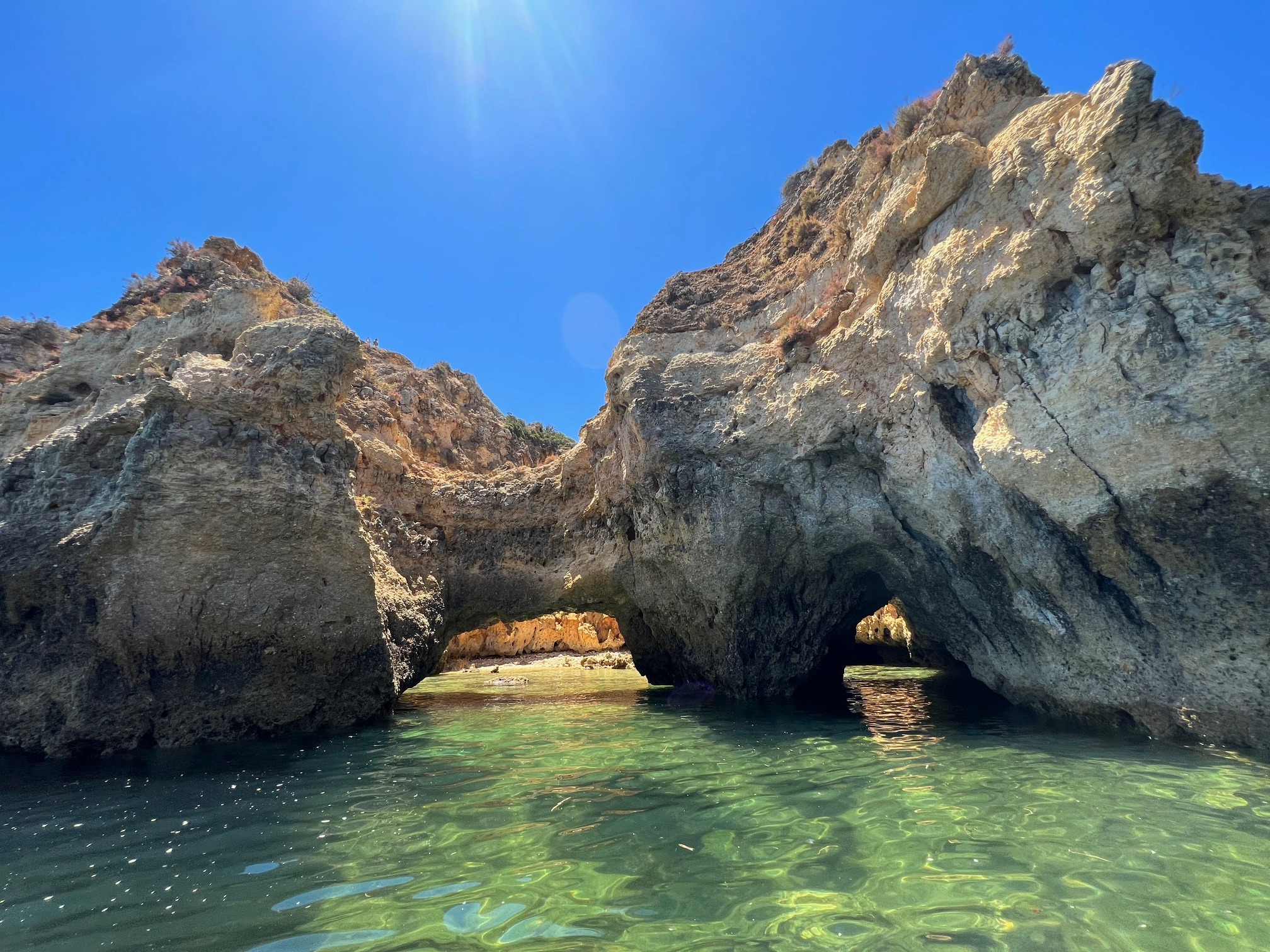 The image showcases a natural rock formation with an arch over clear blue water under a bright sky.