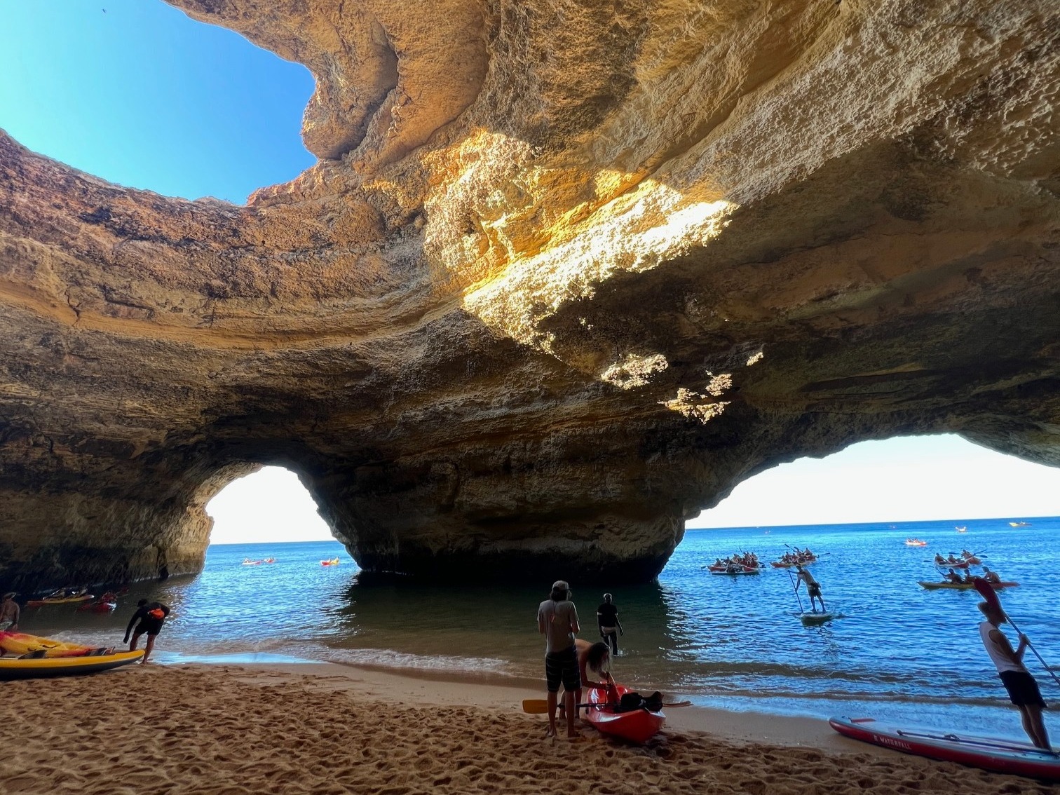 The image shows a natural cave with sunlight streaming through the top, water, and boats on one side, and visitors exploring the rugged interior.