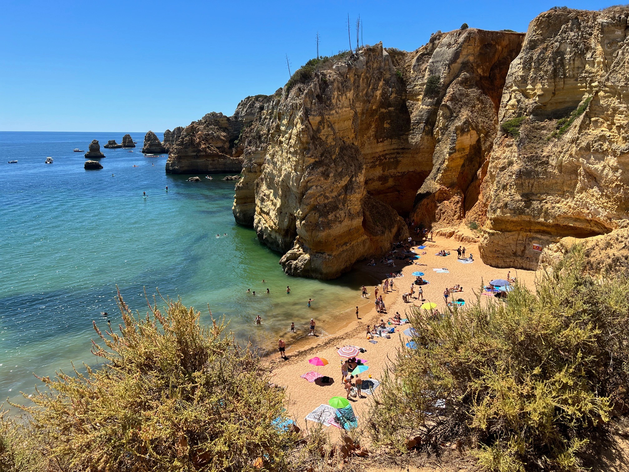 The image depicts a scenic beach with golden cliffs, clear blue water and people enjoying the sun under colorful umbrellas.