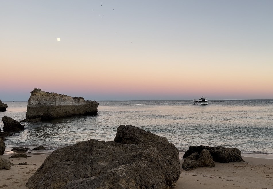 The image portrays a peaceful beach scene at dusk with a boat on the water, large rocks on the sand, and the moon visible in the sky.
