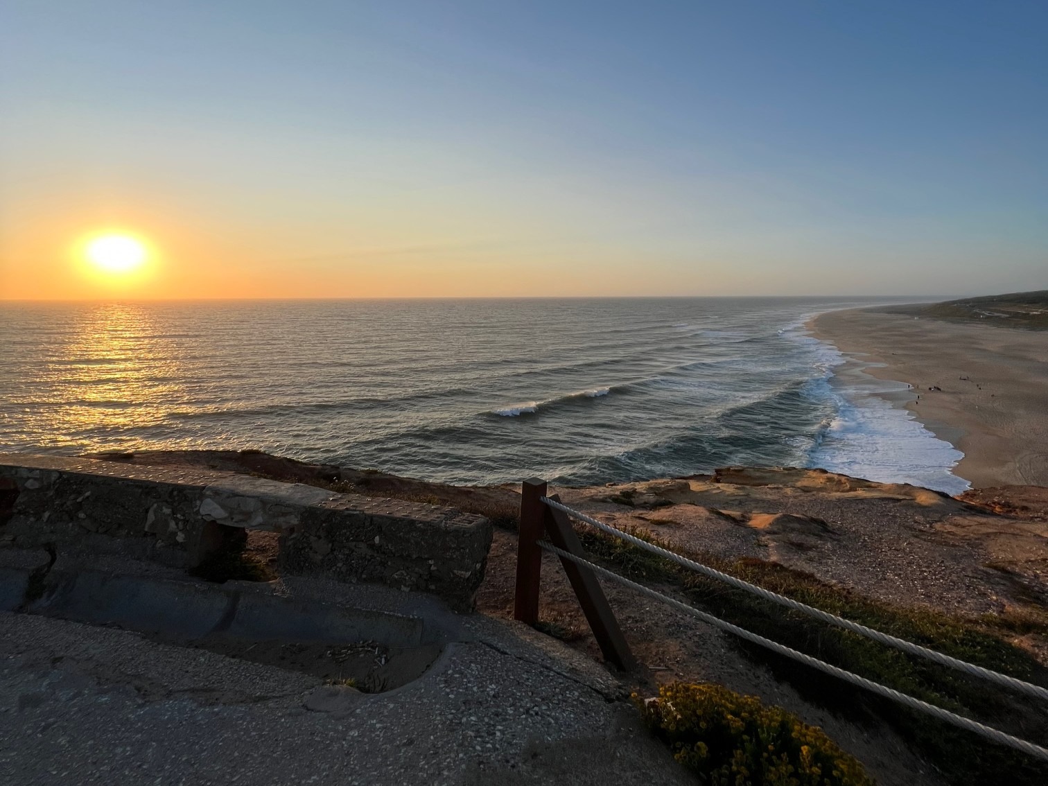 The image captures a coastal sunset with the sun casting a warm glow over the sea and beach, and a railing leading towards the water.