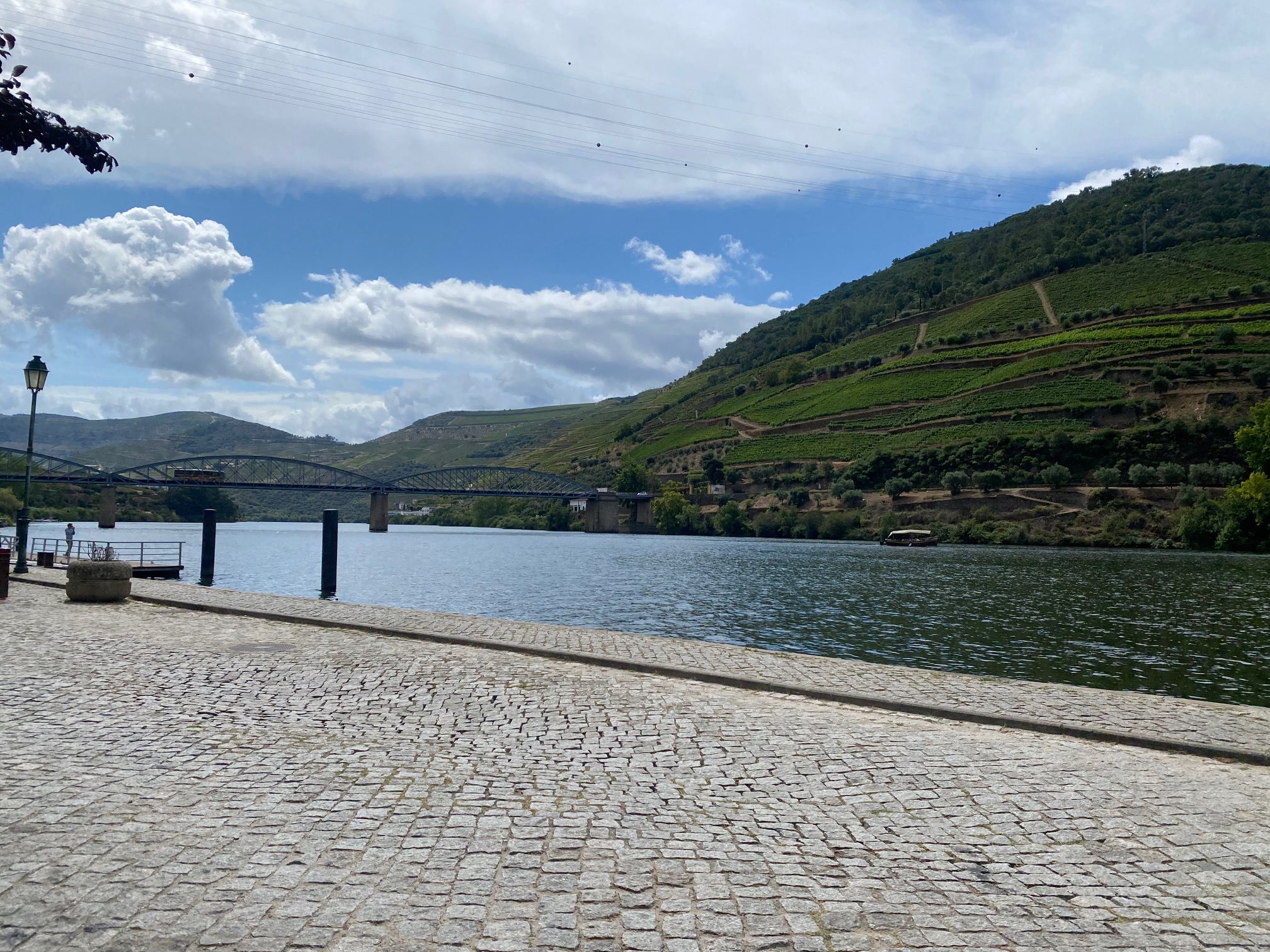 The image displays a riverside view with a cobblestone promenade, a bridge in the distance, and terraced hills under a partly cloudy sky.