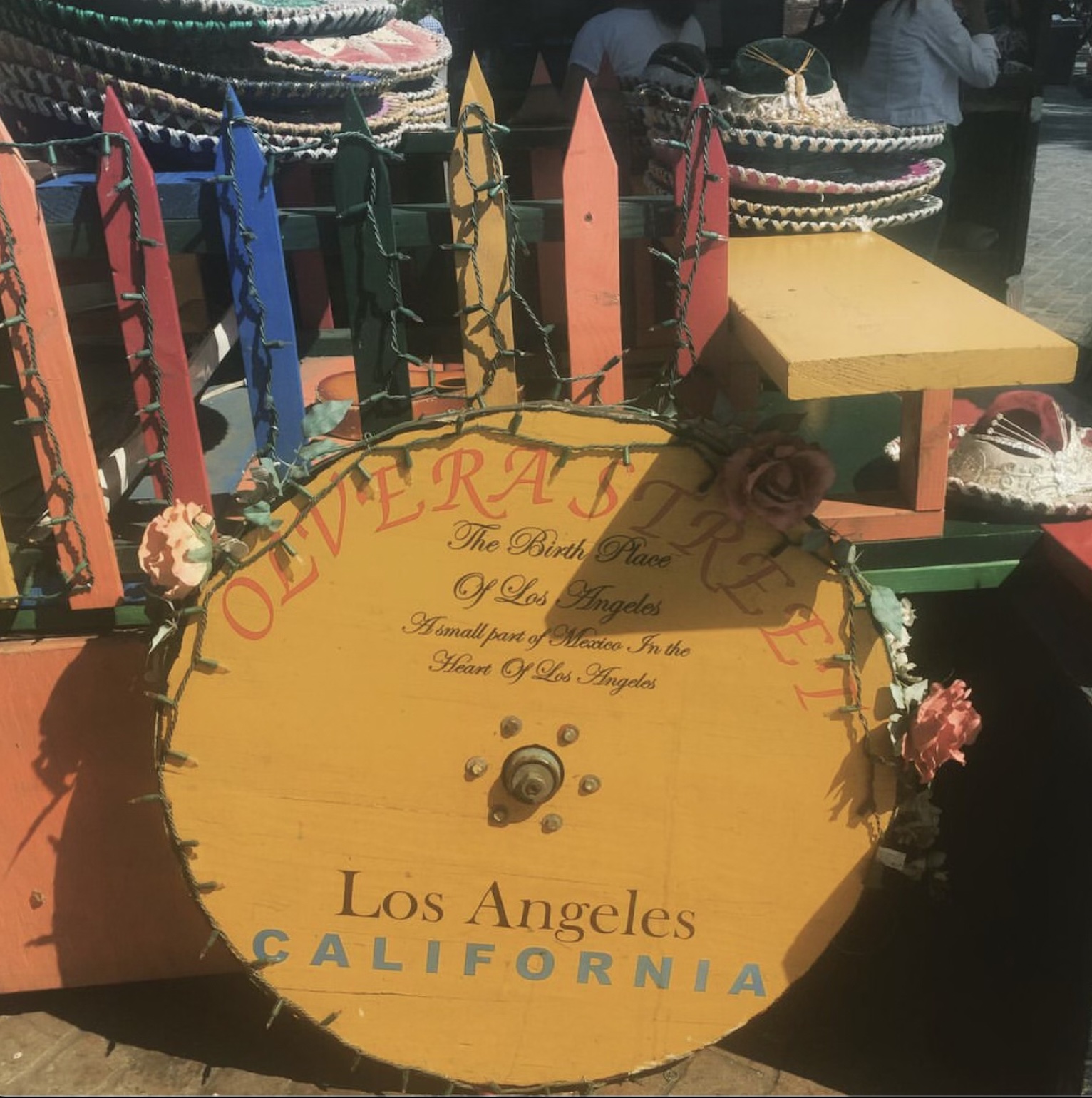 A yellow circular wooden sign signaling Olvera Street in Los Angeles with a colorful fence behind