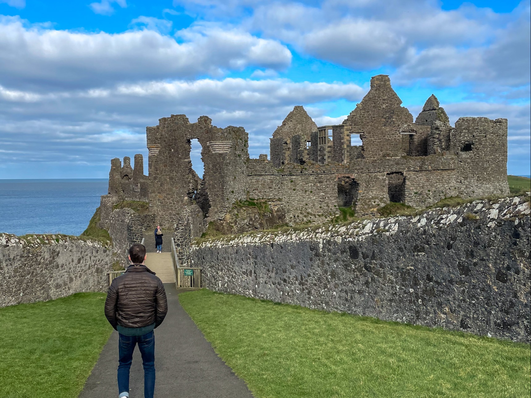 A person approaches the ruins of an ancient stone castle, with the ocean as a backdrop