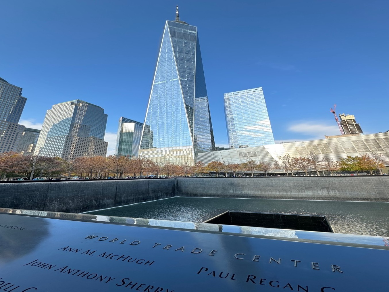 The image captures the 9/11 Memorial, with names inscribed and skyscrapers under a clear sky, marked by the text “WORLD TRADE CENTER.”
