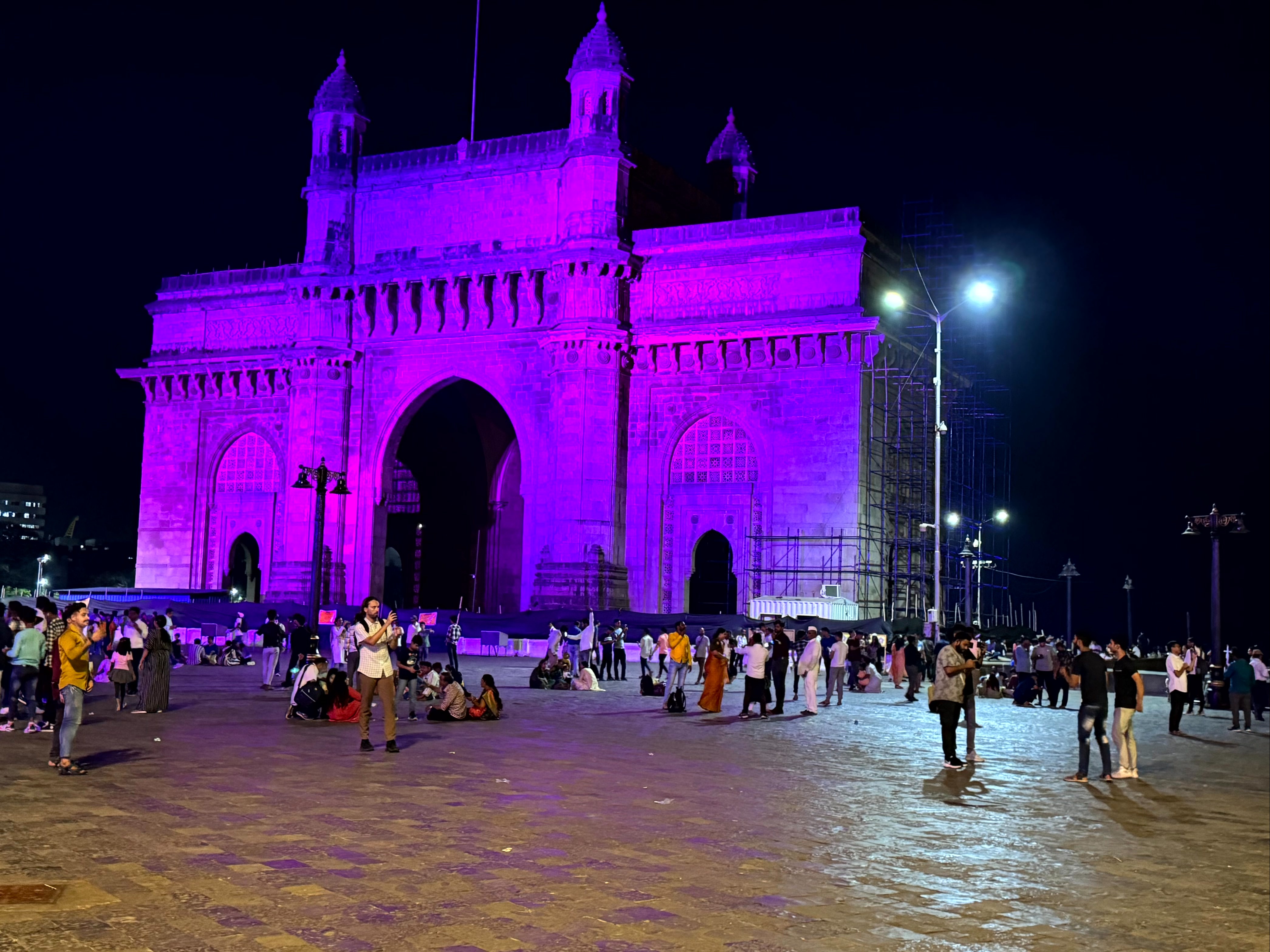 The iconic Gateway of India monument, bathed in purple light at night, with a lively crowd in the foreground.
