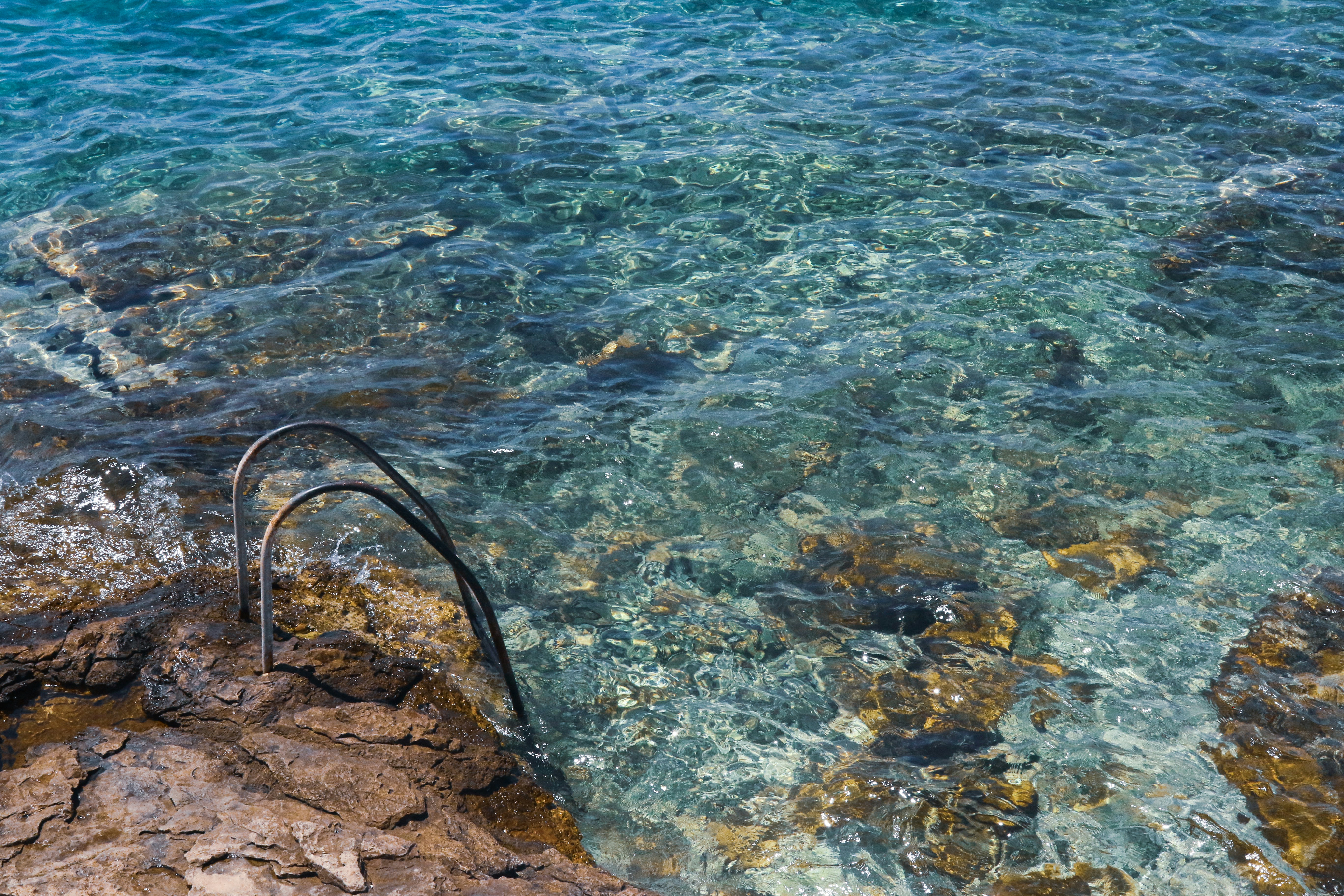 A small, humble, metal ladder embedded into the rocky coastline and descending into the crystal clear water of the Adriatic Sea.