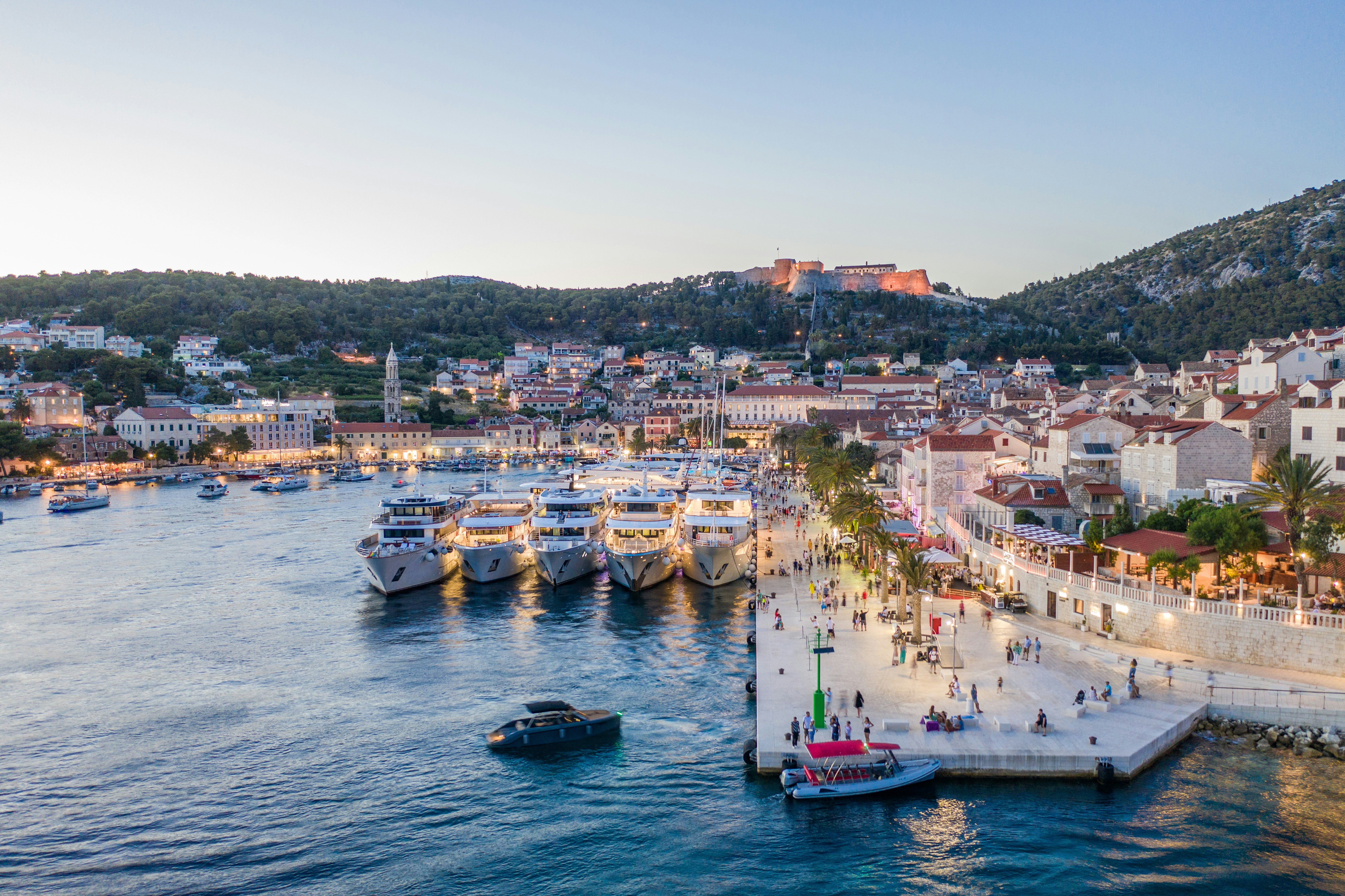 A view of Hvar's marina with boats and small ships, shops, restaurants and people enjoying the site at dusk.