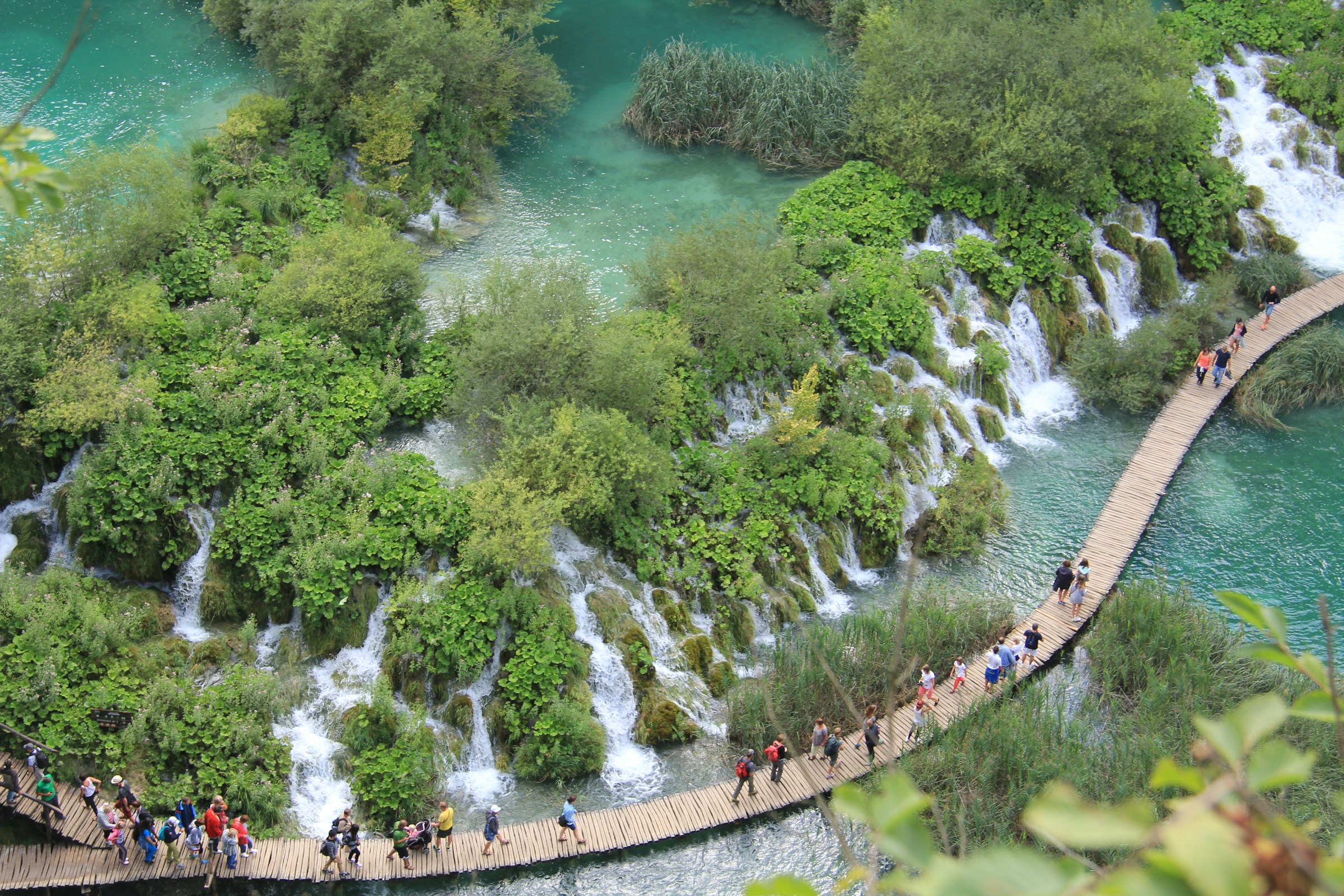A view from above of people walking down a curvy, wooden, foot bridge over blue water, surrounded by mini waterfalls, trees and natural greenery at Plitvice Lakes National Park