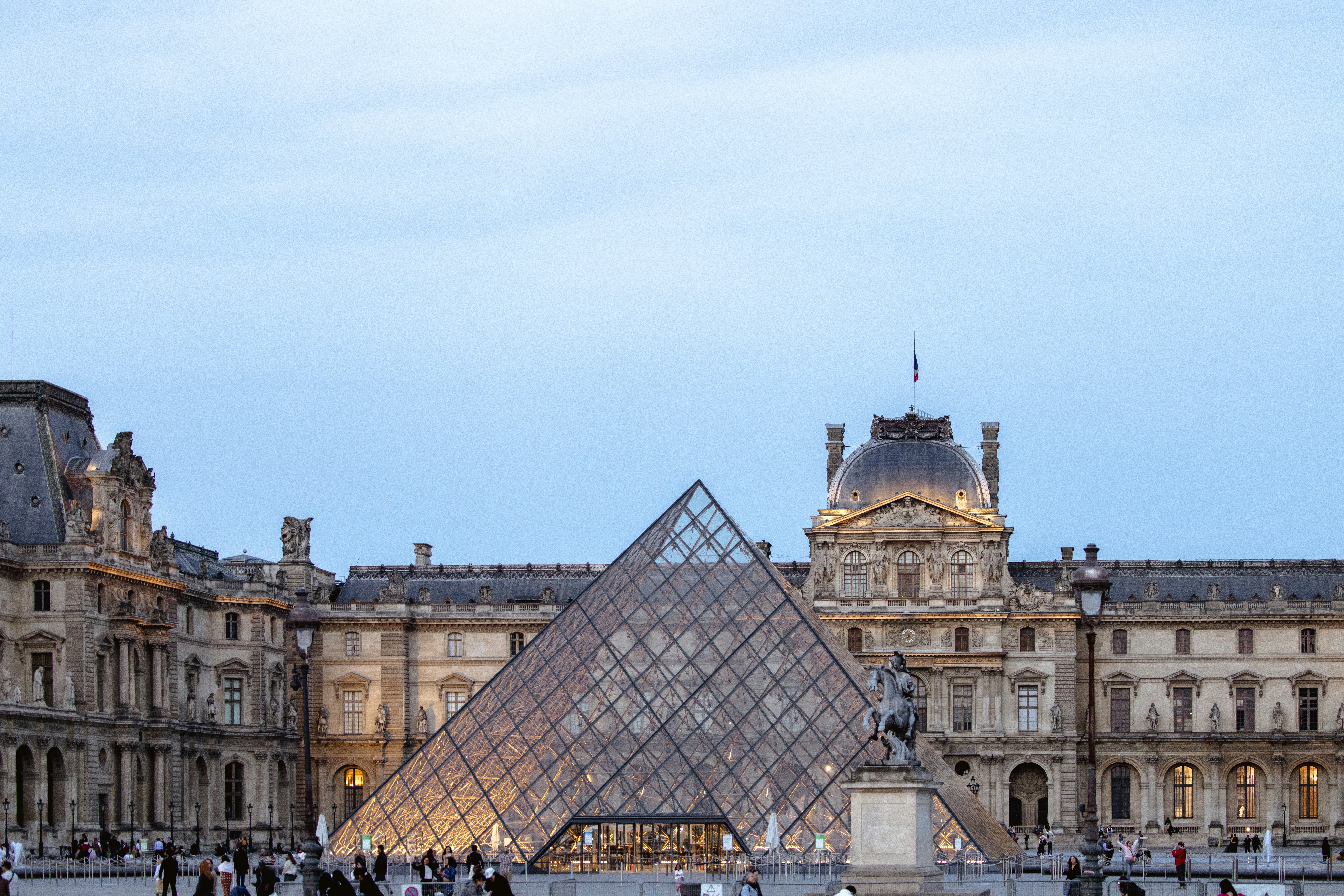 The image captures the iconic Louvre Pyramid illuminated in the early evening time.