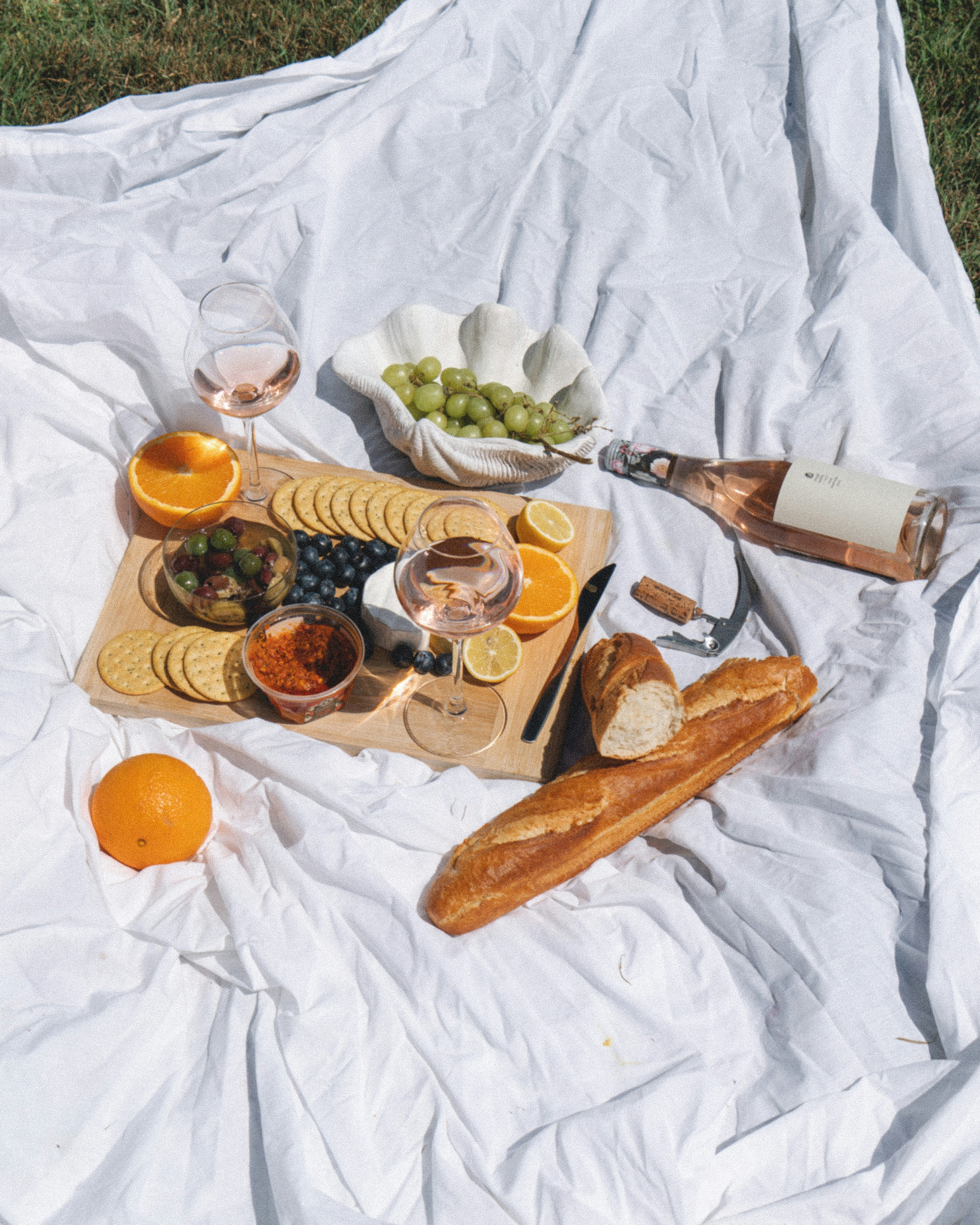 A white blanket with a picnic of cheese, crackers, wine and fruits laid out on top of it.