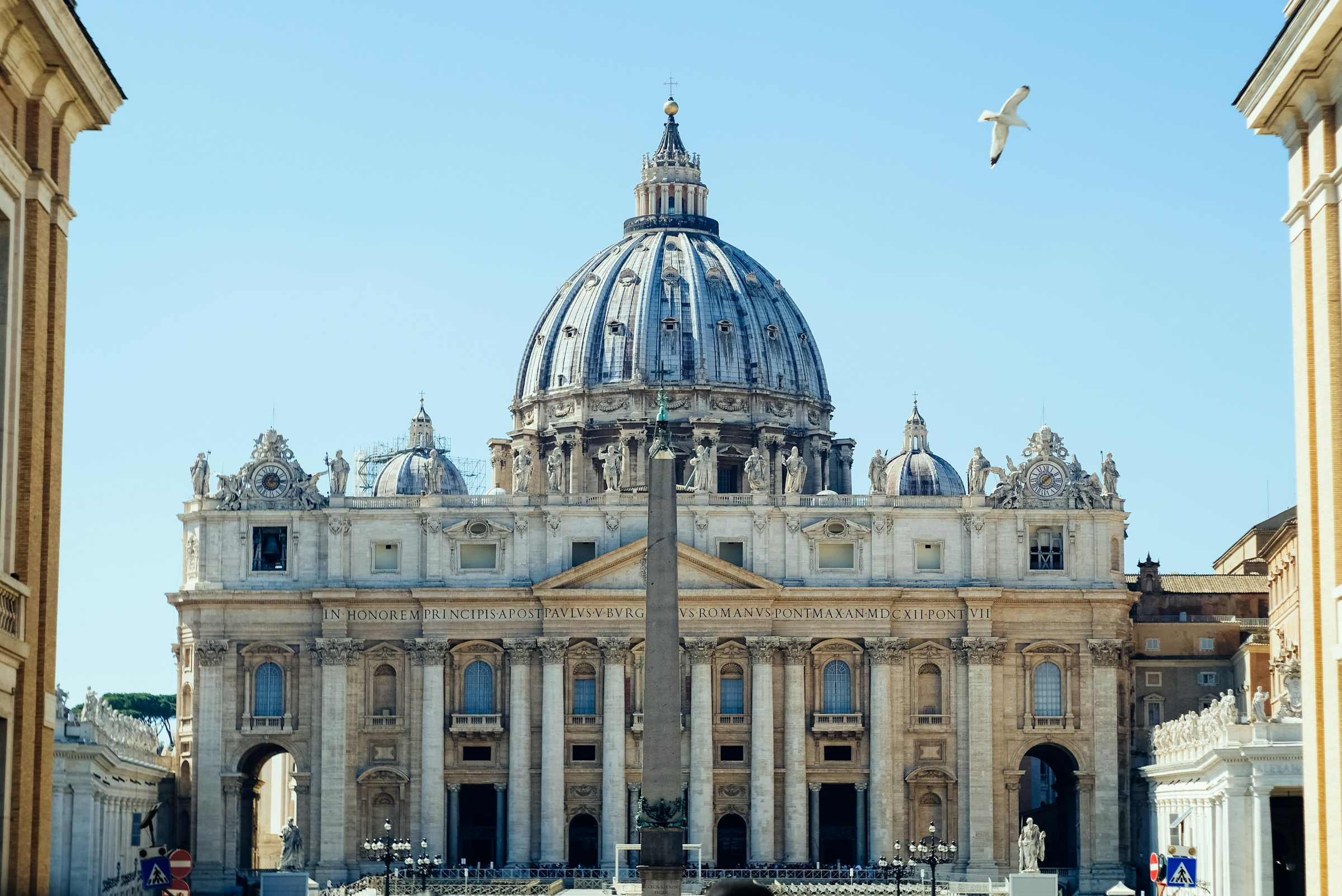 St. Peter's Basilica, an ornate church with stone pillars and a blue dome, during daytime with birds flying above.